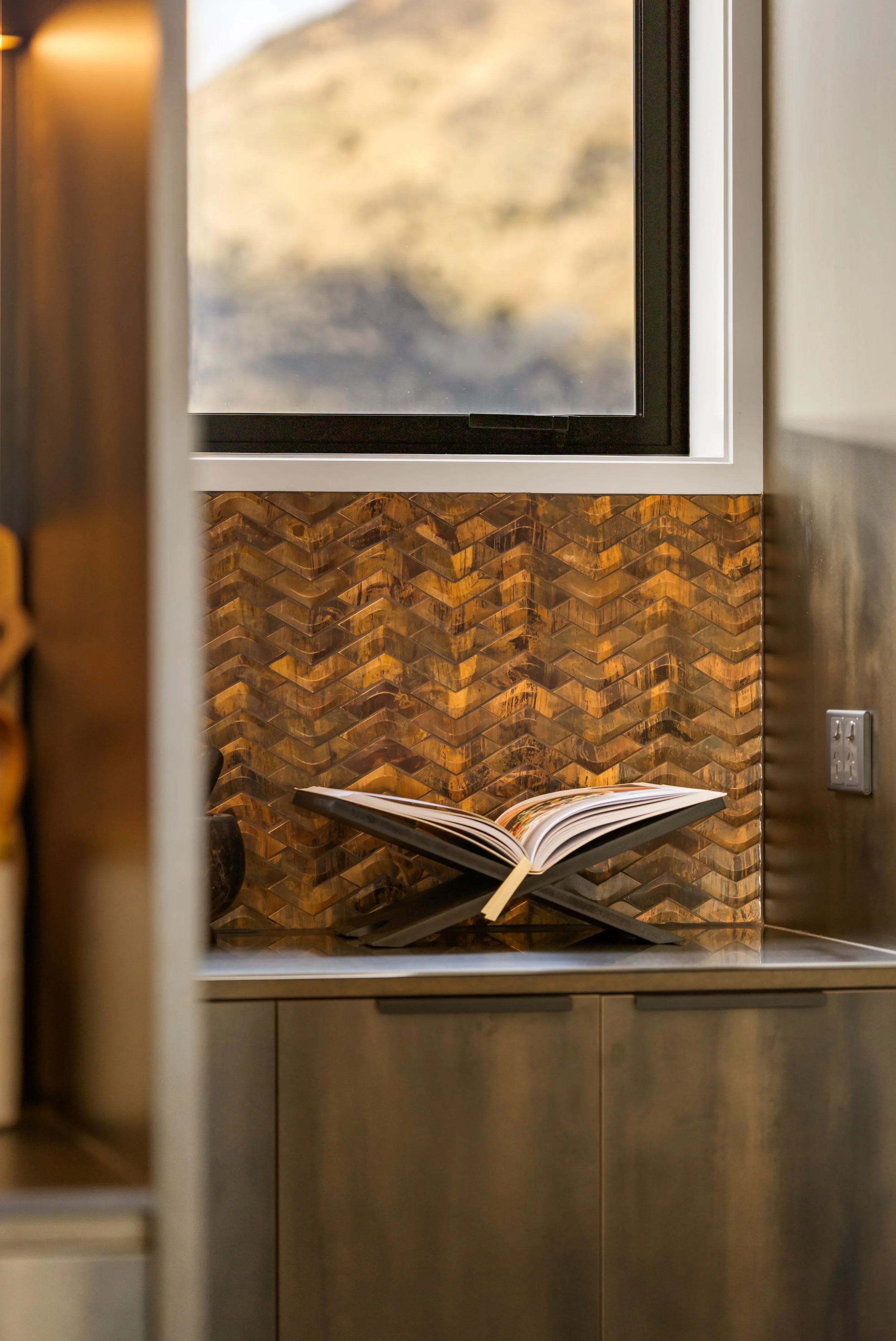 Open book on a black metal stand placed on a kitchen counter with bronze mosaic tiles and a window showing a mountain landscape in the background.