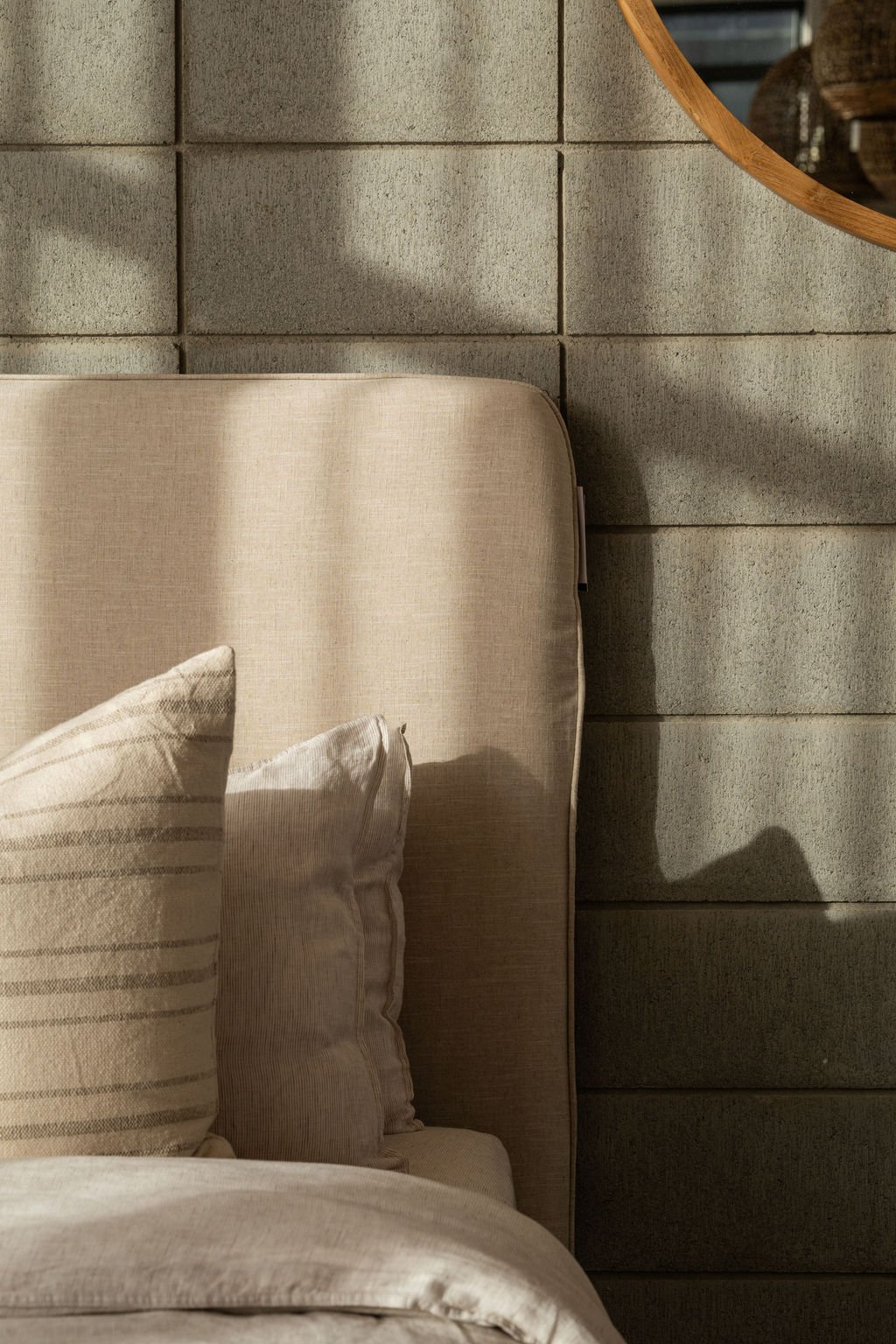 Close-up of a beige upholstered headboard, with beige and cream pillows resting against it, beside a gray stone wall and a round wooden mirror in the upper right corner.