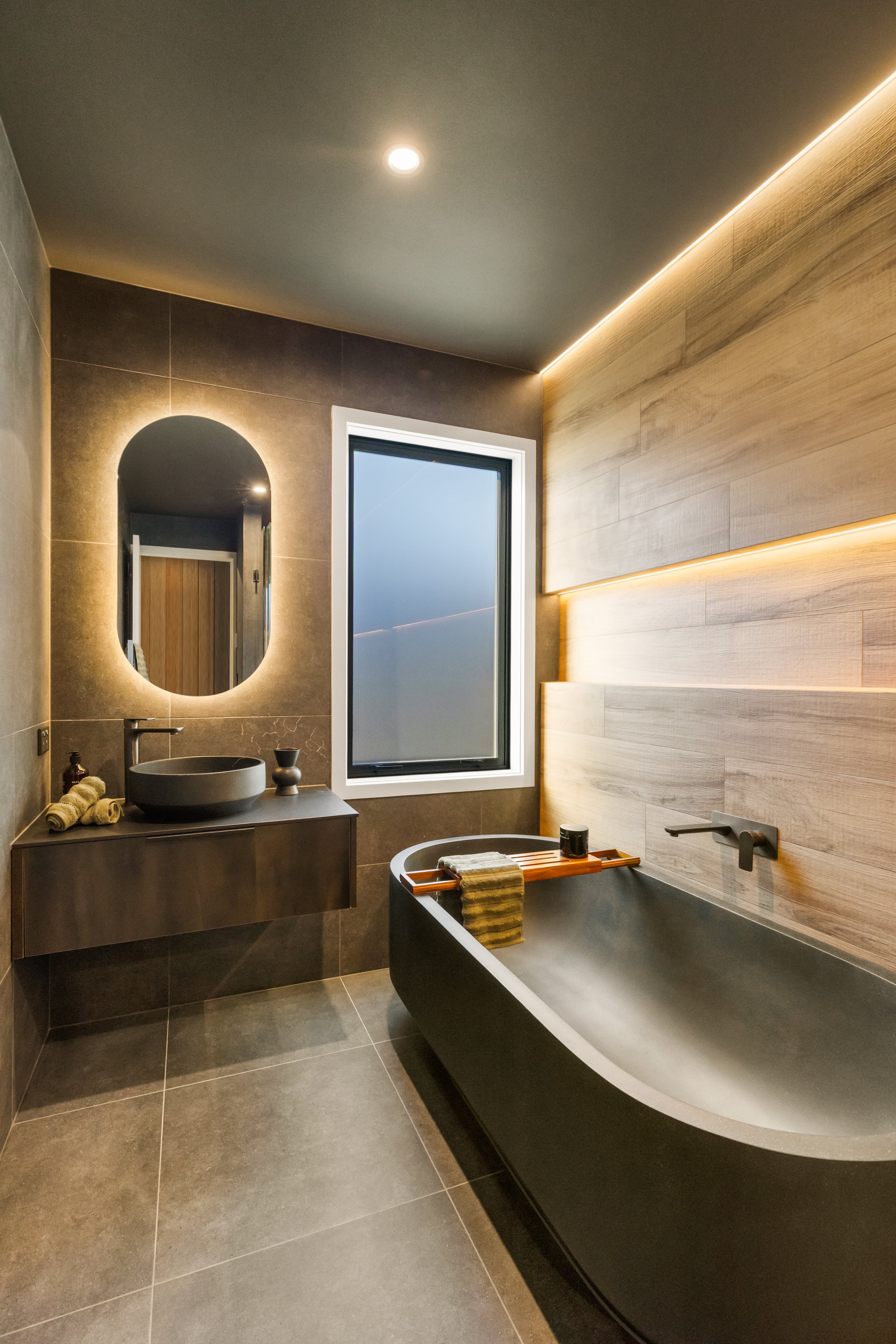 Modern bathroom featuring a black bathtub, wooden wall panels, a gray vanity with a vessel sink, a circular mirror with backlighting, a window, and subtle ceiling lighting.