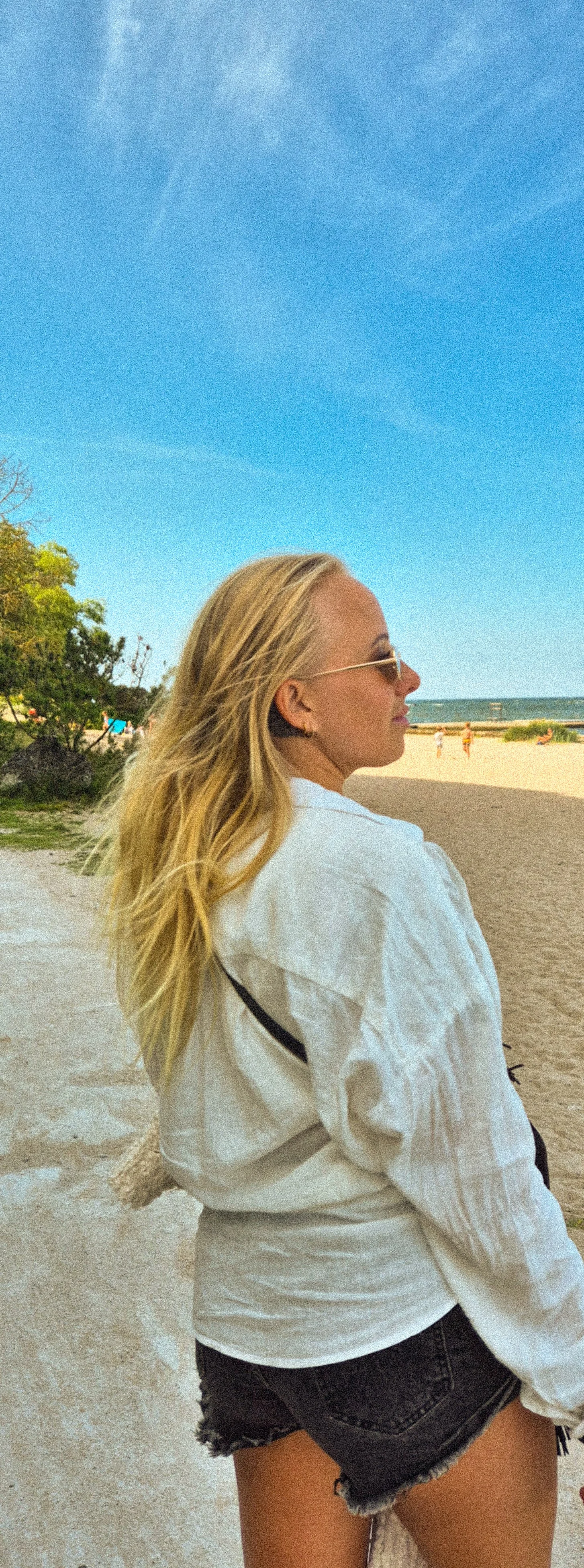 Woman with long blonde hair wearing sunglasses, white shirt, and black shorts, standing on a beach under a blue sky.