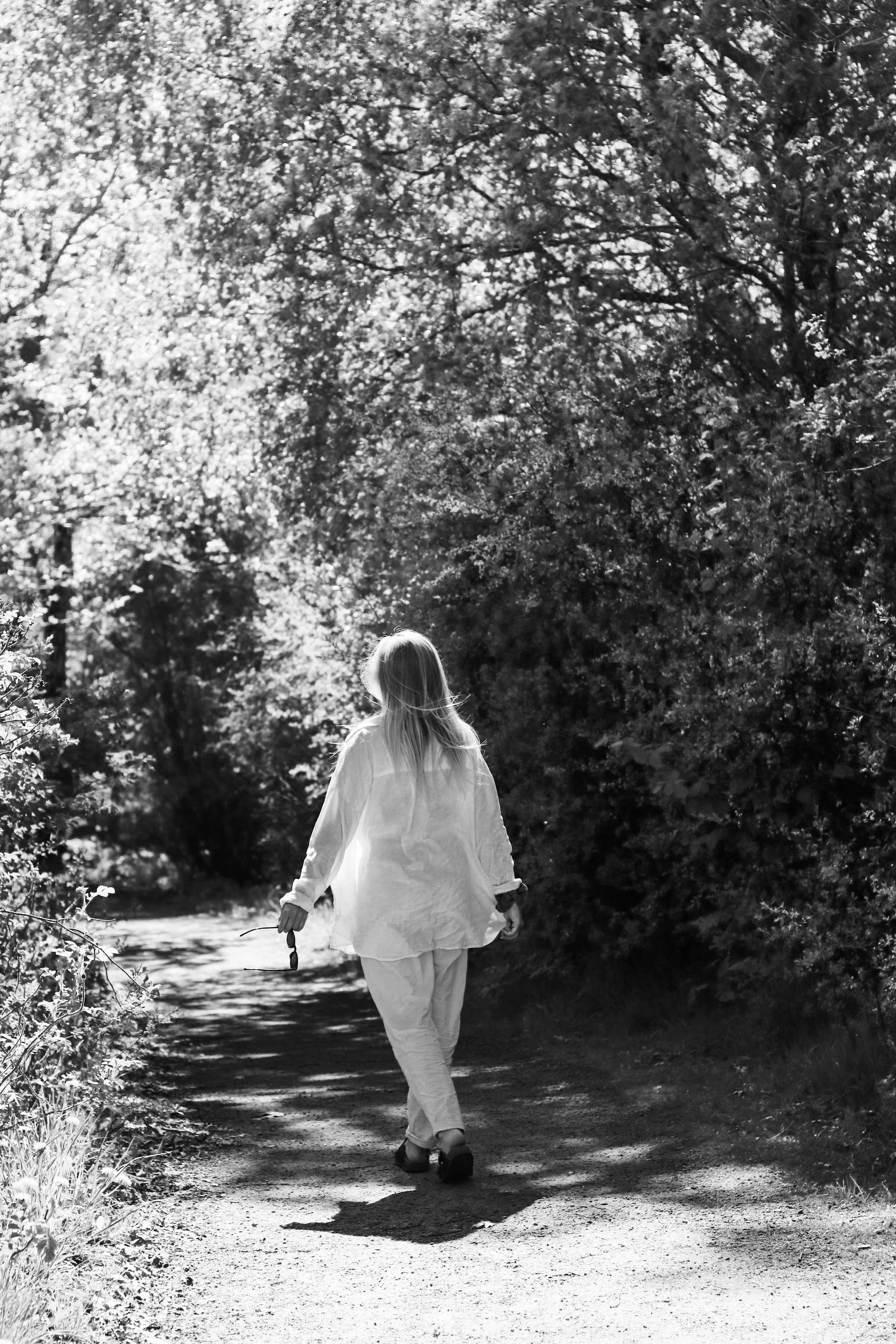 A woman walking alone on a wooded dirt path surrounded by trees in sunlight, captured in black and white.