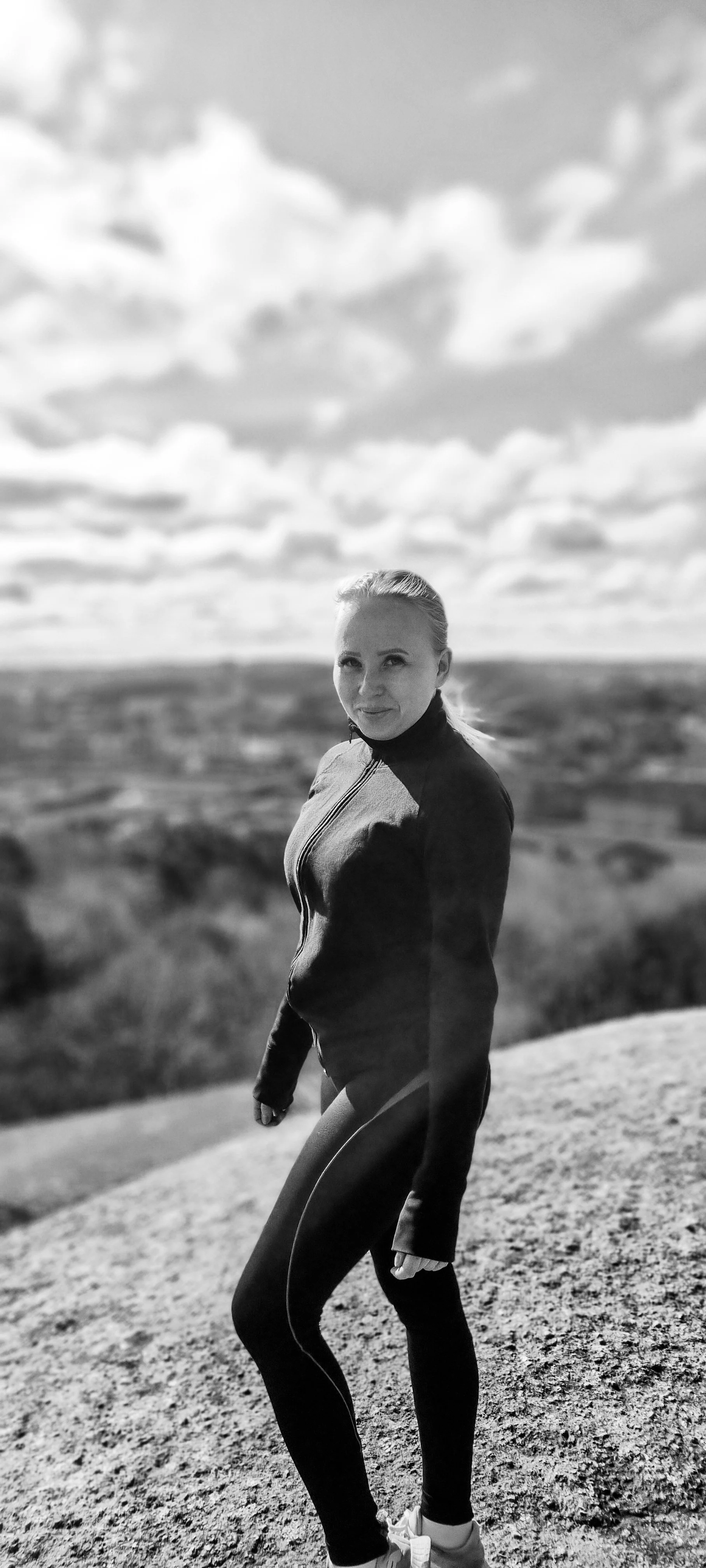 Black and white photo of a woman standing outdoors on a rocky surface with a landscape and cloudy sky in the background.