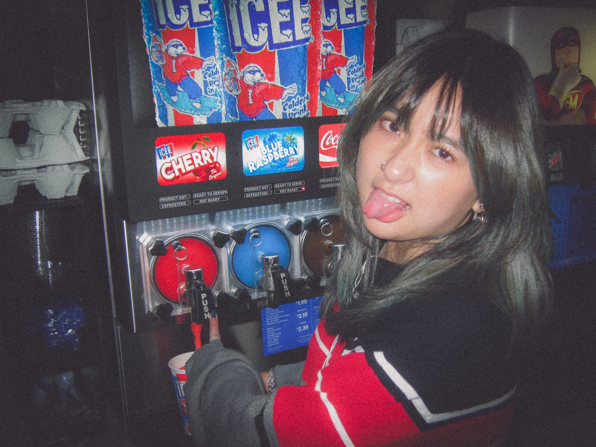 A young woman with dark hair and a nose piercing sticking out her tongue while at a soda fountain, with various sodas and ice pops in the background.