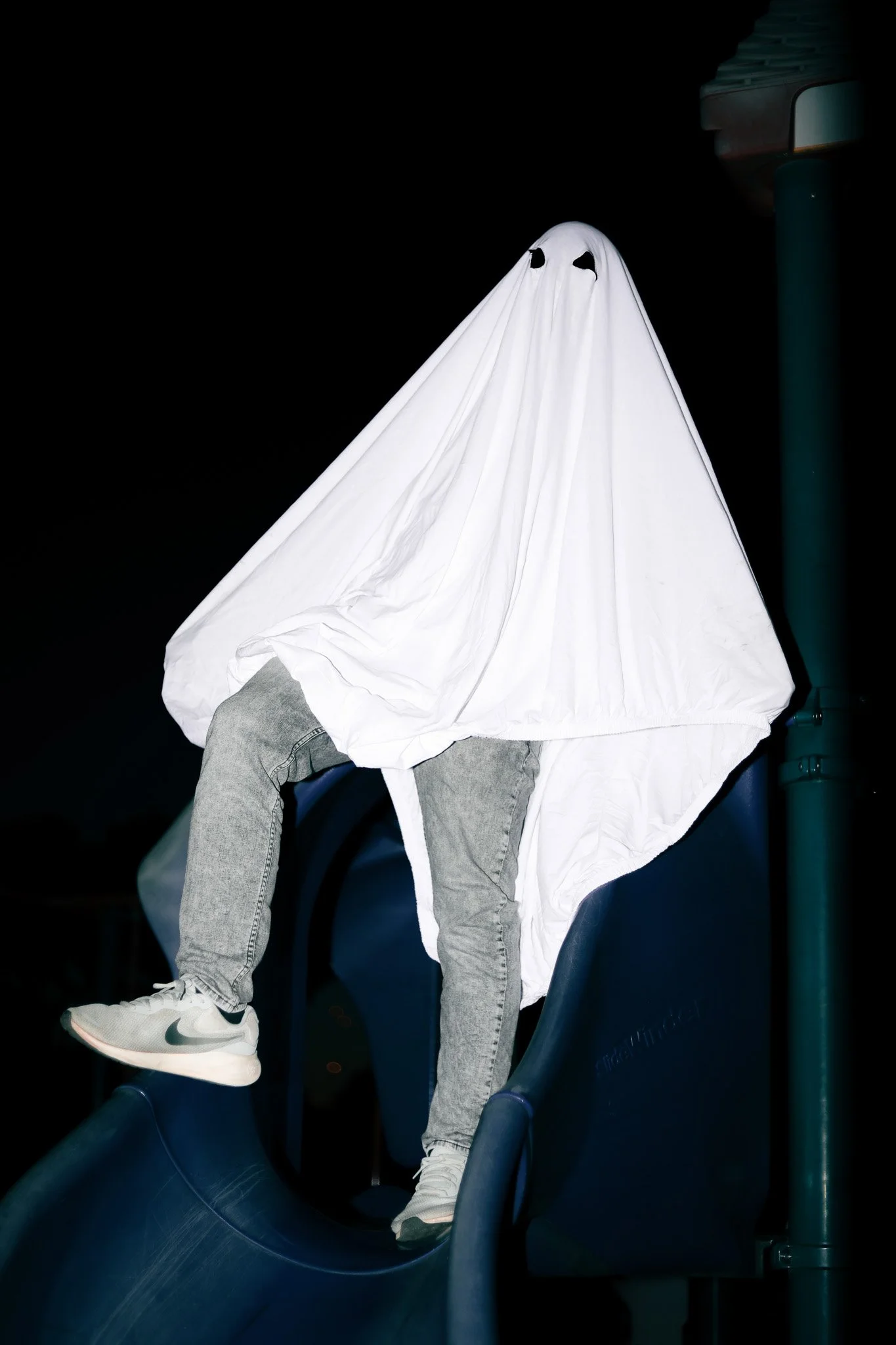 Person sitting on a blue stadium seat with a white sheet draped over their head, resembling a ghost, at night.