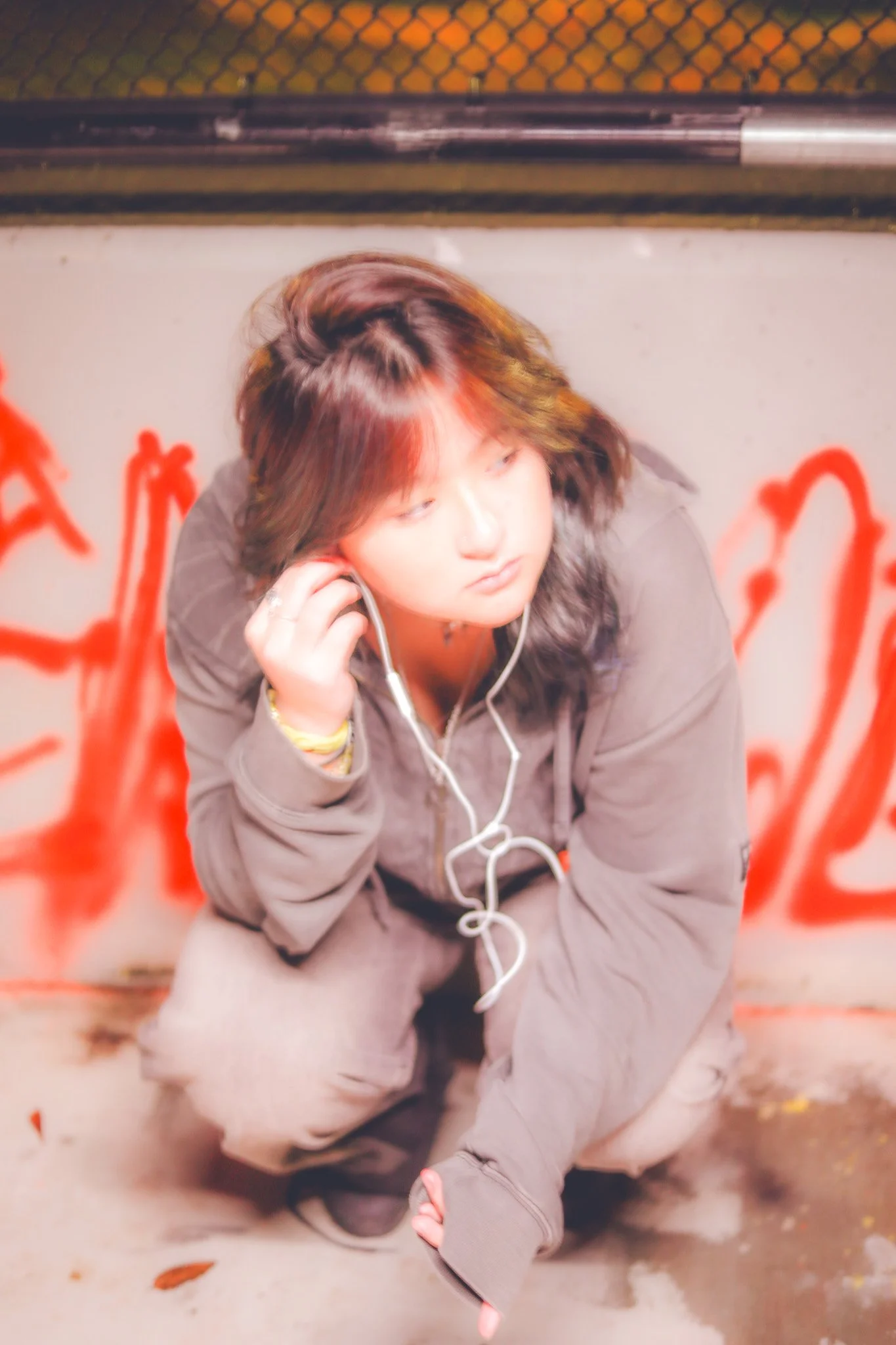 A person wearing a gray jacket, squatting on a sidewalk with graffiti in the background, listening to headphones.