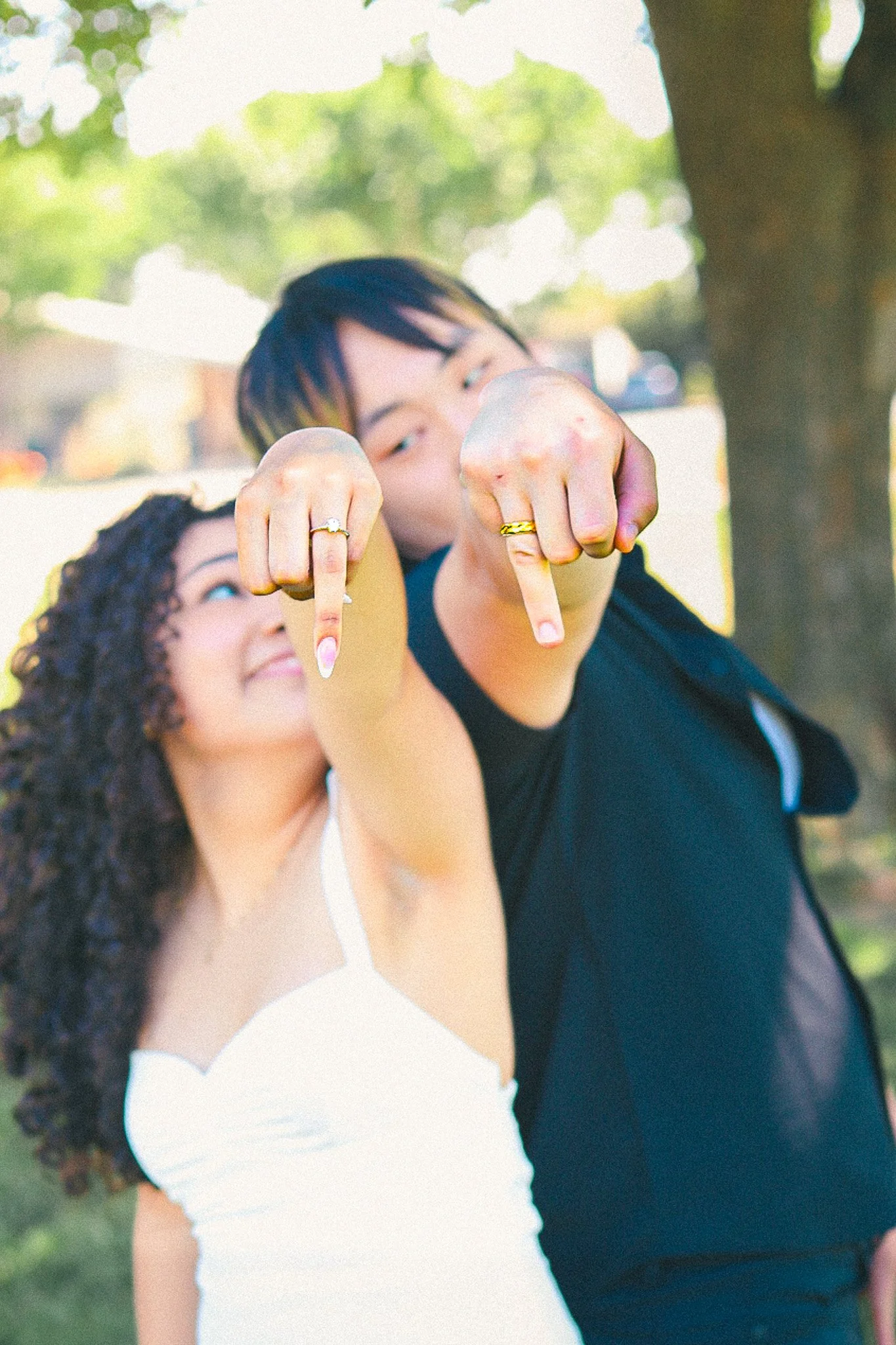 Two people showing their rings while pointing at the camera outdoors