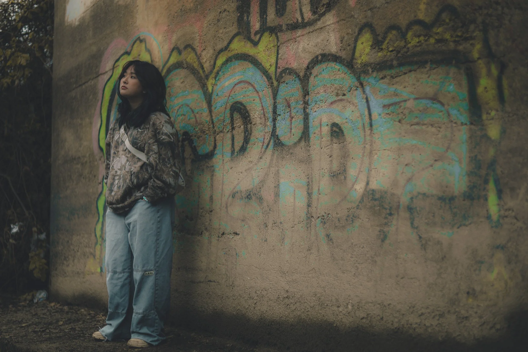 A young woman standing against a graffiti-covered concrete wall outdoors.