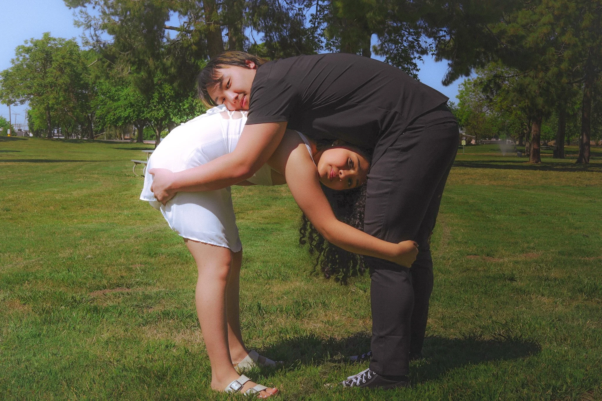 Two women, one in white and one in black, are performing a coordinated yoga pose in a park with green grass and trees in the background.