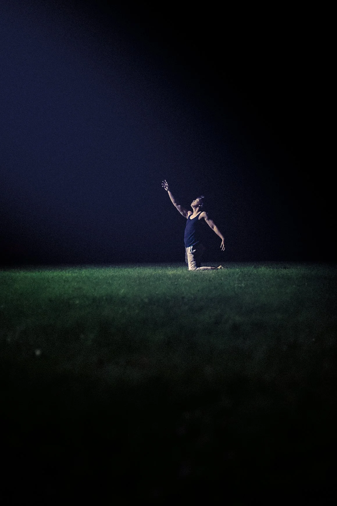 Person kneeling on grass at night with one arm reaching upward, illuminated by a focused light.
