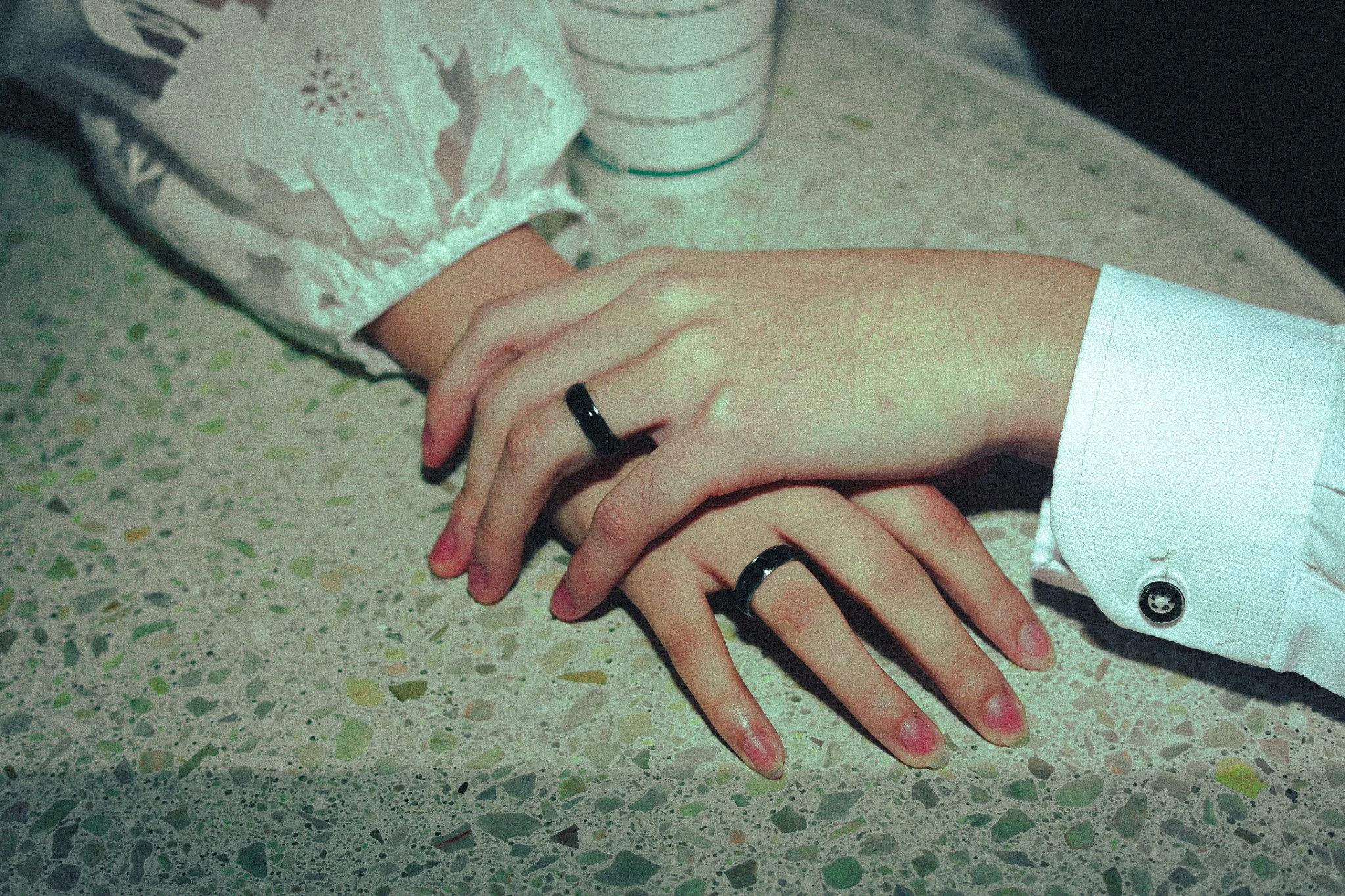 Two people, one with a light skin tone and the other with a darker skin tone, holding hands on a speckled countertop. The person with the darker skin tone wears a ring on their index finger, and the person with the lighter skin tone wears a ring on t