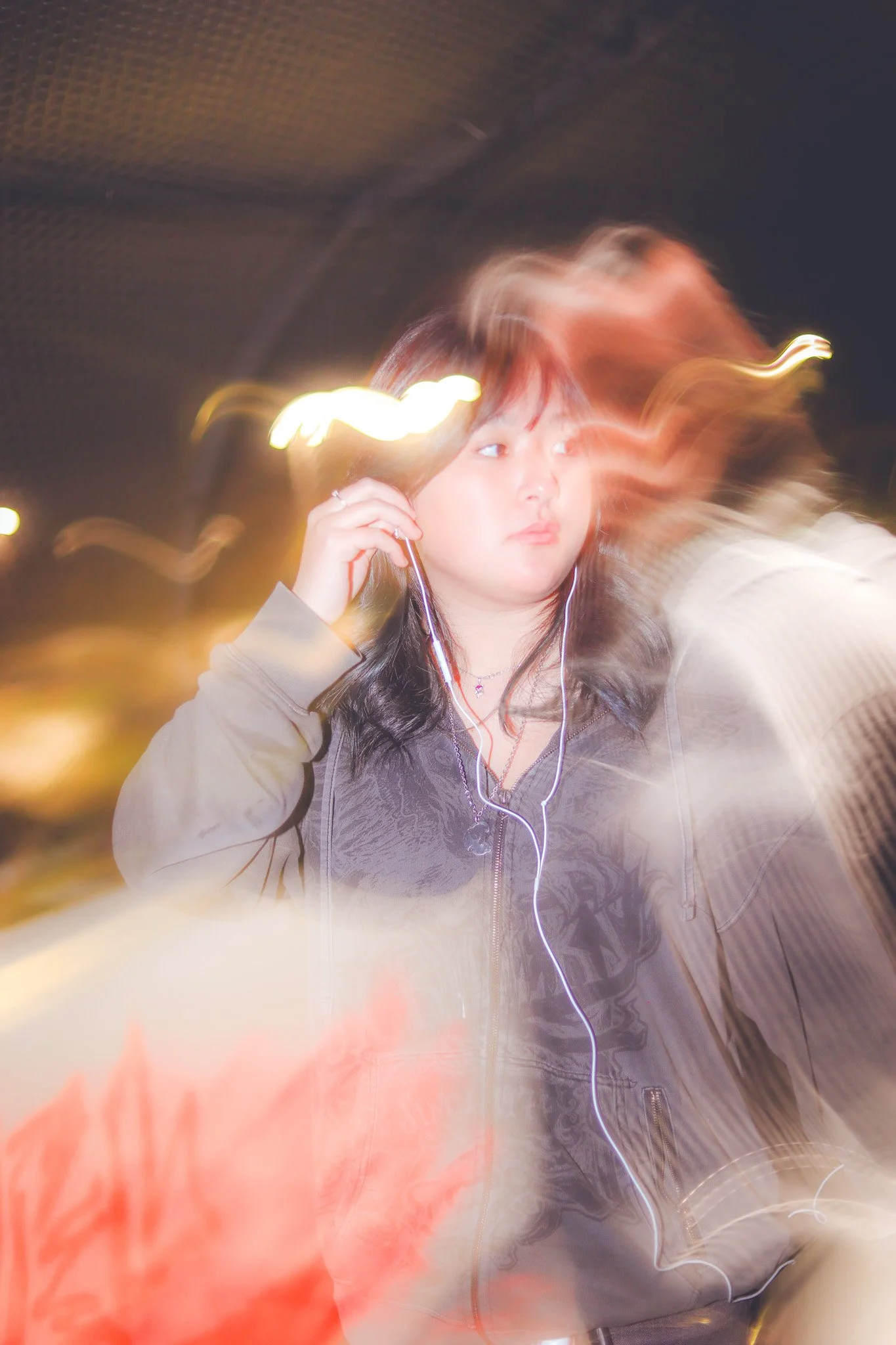 A young woman with dark hair wearing a gray jacket, listening to music with white earbuds, standing outdoors at night with light streaks around her due to camera movement.