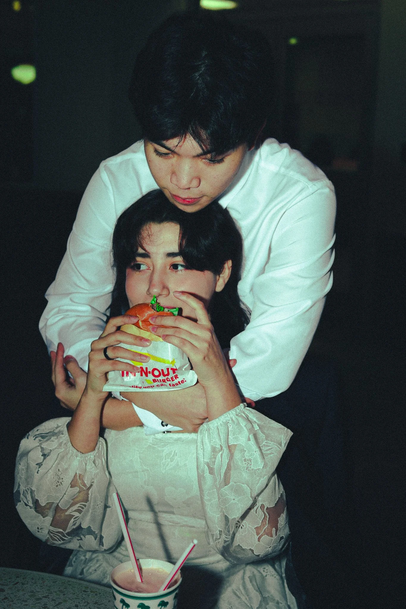 Two people sharing a burger at a table with a pink drink. One person holds the burger and the other embraces them from behind.