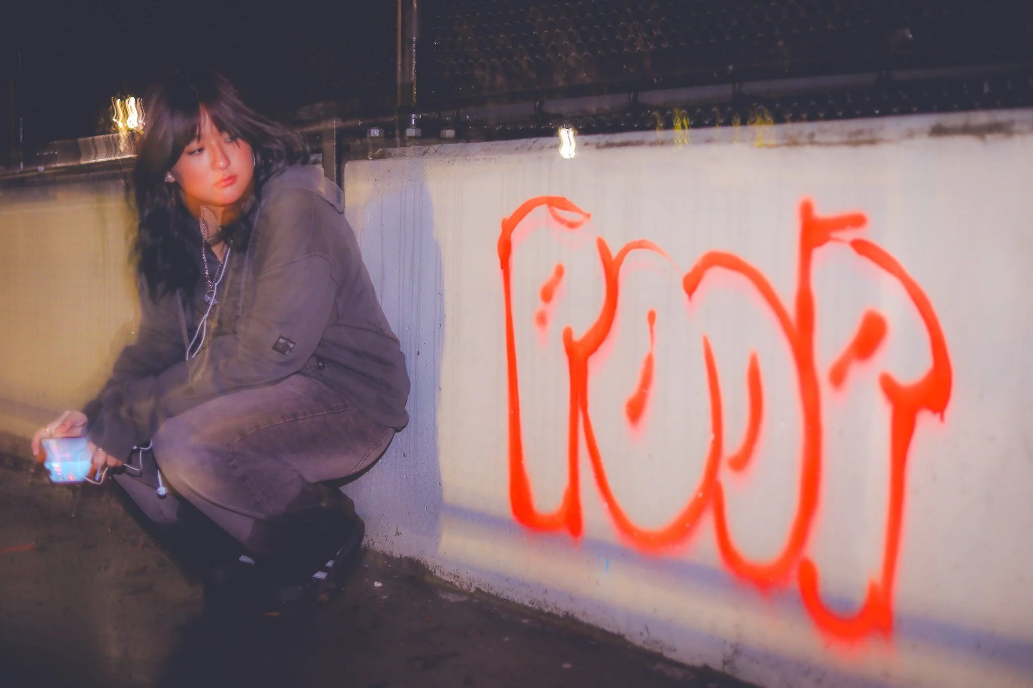 A young woman with black hair squatting near a graffiti-covered wall at night, holding a smartphone, with the word 'LOVE' spray-painted in red on the wall.