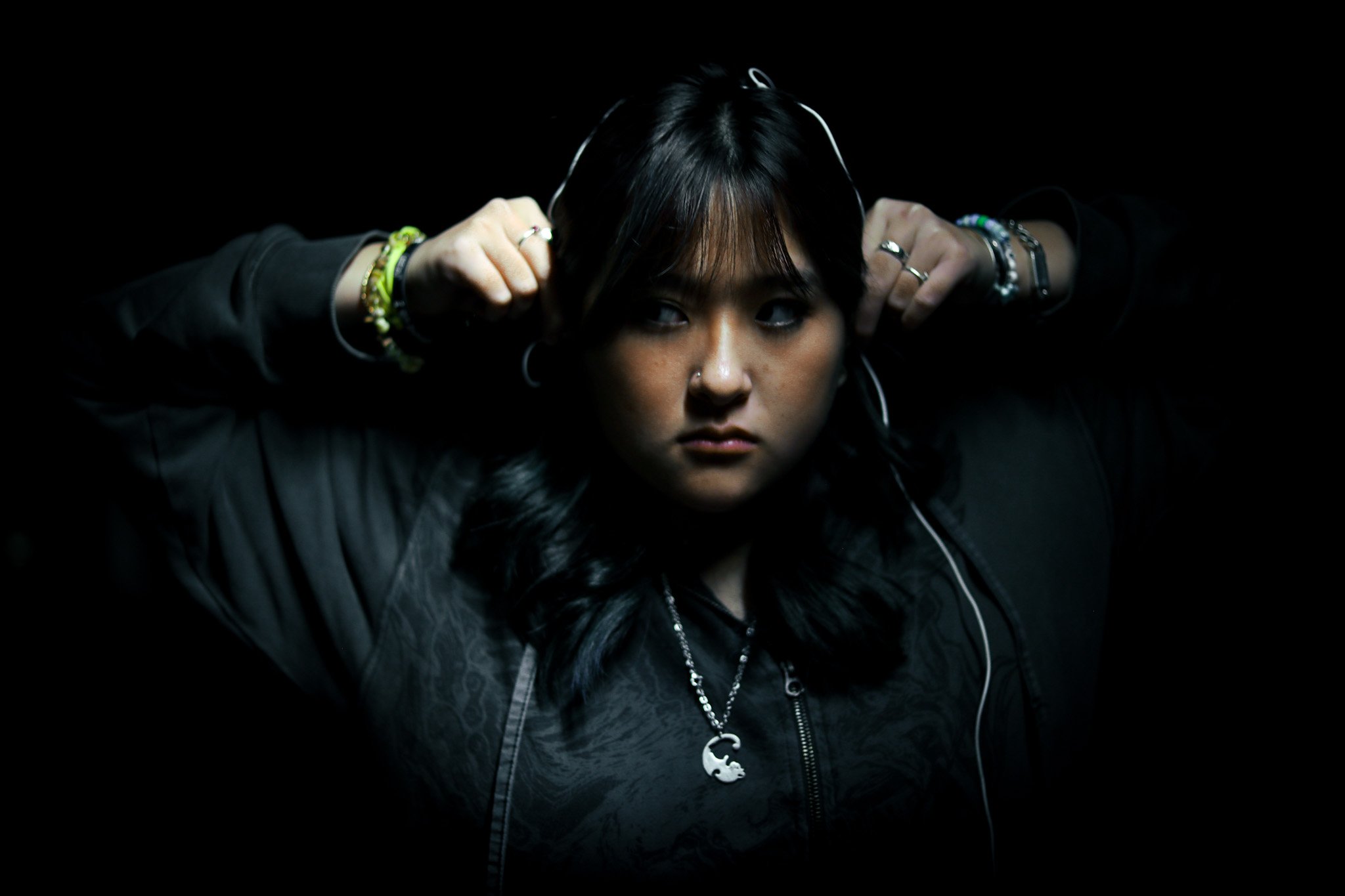 A young woman with dark hair and multiple bracelets, wearing a black jacket and headphones, adjusting the headphones against a black background.