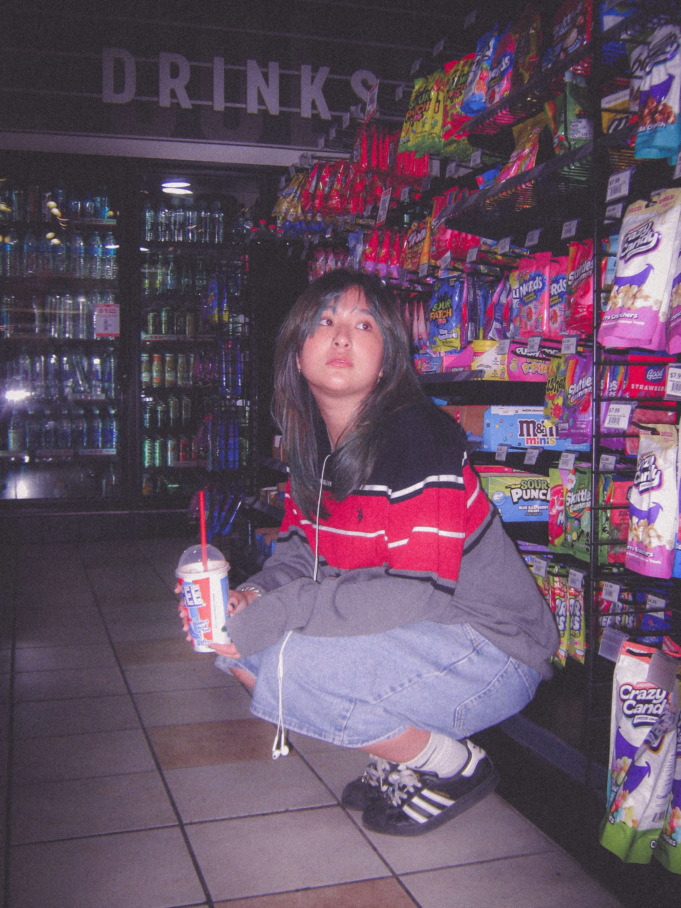 Young woman crouching in a convenience store aisle holding a beverage, surrounded by snack shelves and a drinks refrigerator in the background.