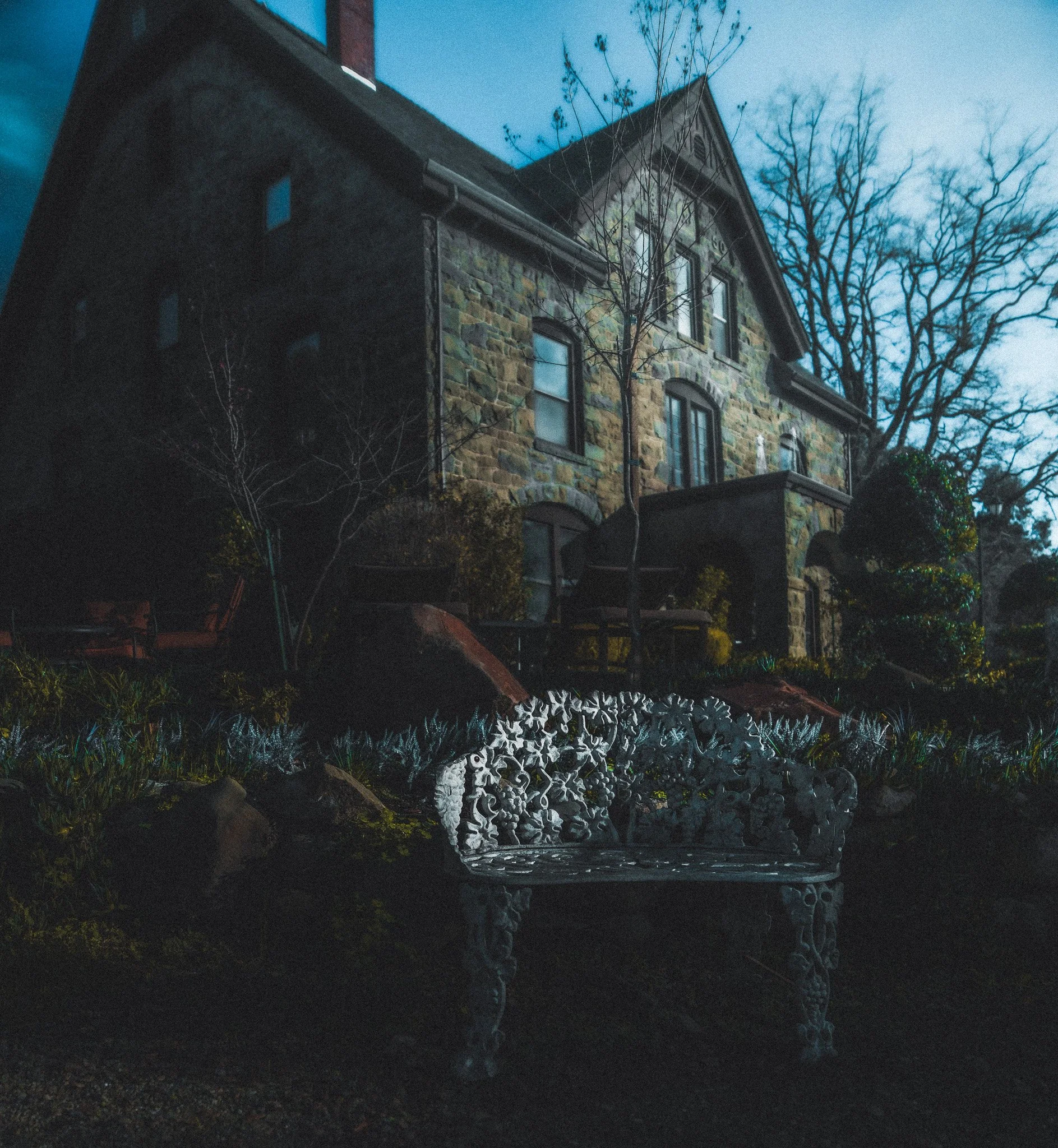 Nighttime view of a stone house with tall trees behind it, and a decorative metal bench in the foreground. The house has multiple windows and a gabled roof.