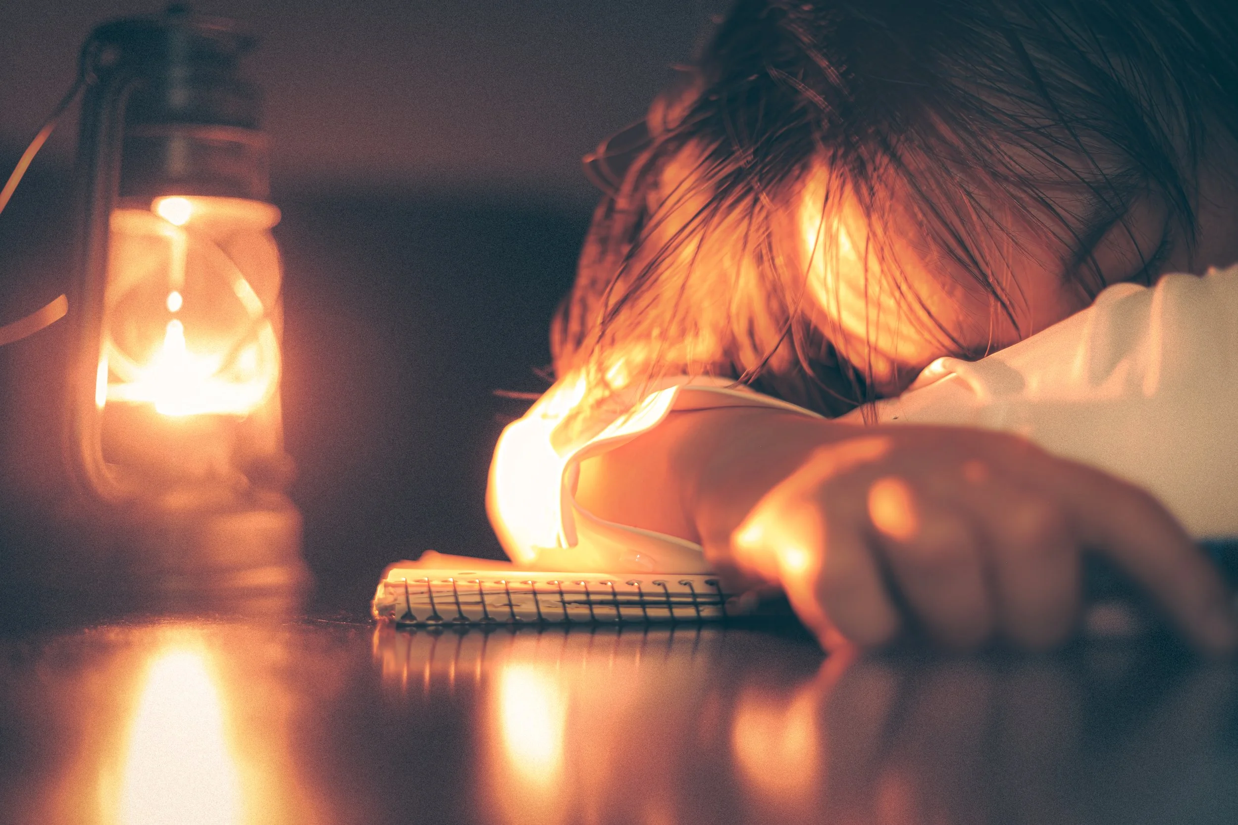 Person with disheveled hair resting face down on a notebook at a desk, illuminated by warm, ambient light from a nearby lantern.