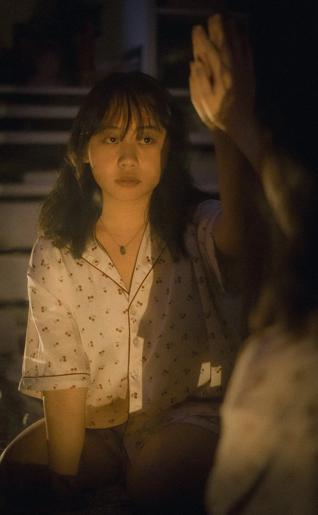 A young woman with dark hair, wearing pajamas with a cherry pattern, is sitting in a dimly lit room, reaching out her hand towards someone or something not visible in the image.