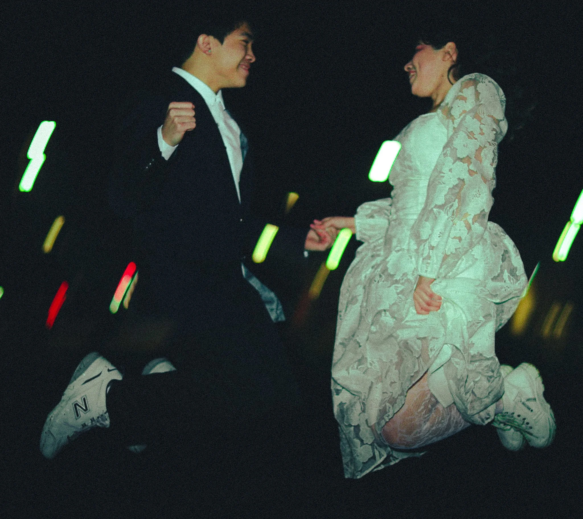 A young man and woman are dancing together at night, with the woman dressed in a patterned dress and the man in a suit, holding hands and smiling.