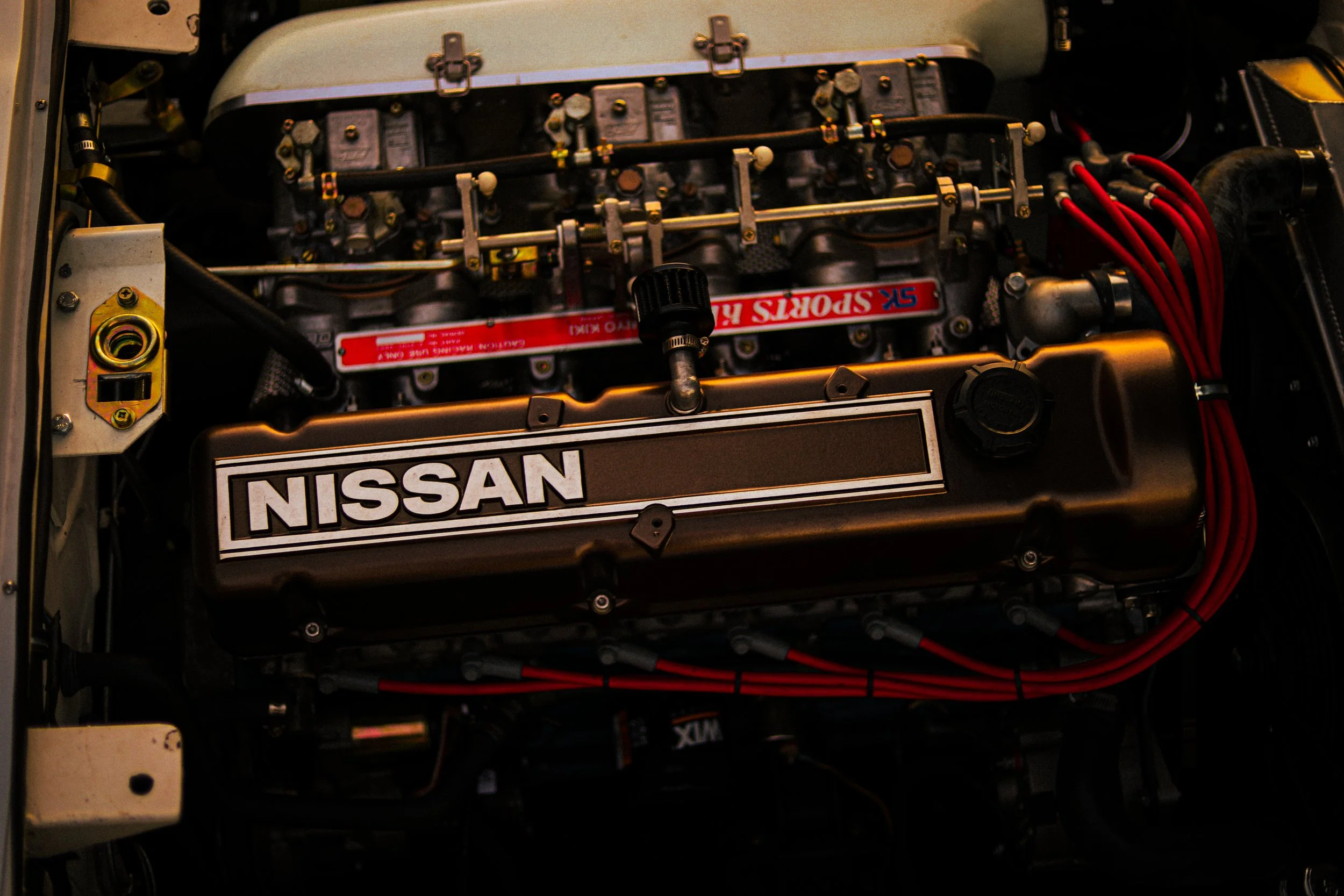 Close-up of a Nissan race car engine with red spark plug wires and a black valve cover labeled 'NISSAN'.