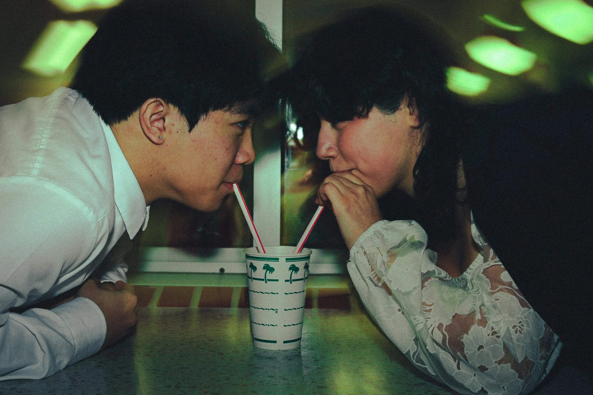 A young man and woman leaning toward each other through a paper cup with straws, sharing a drink, in a retro diner environment.