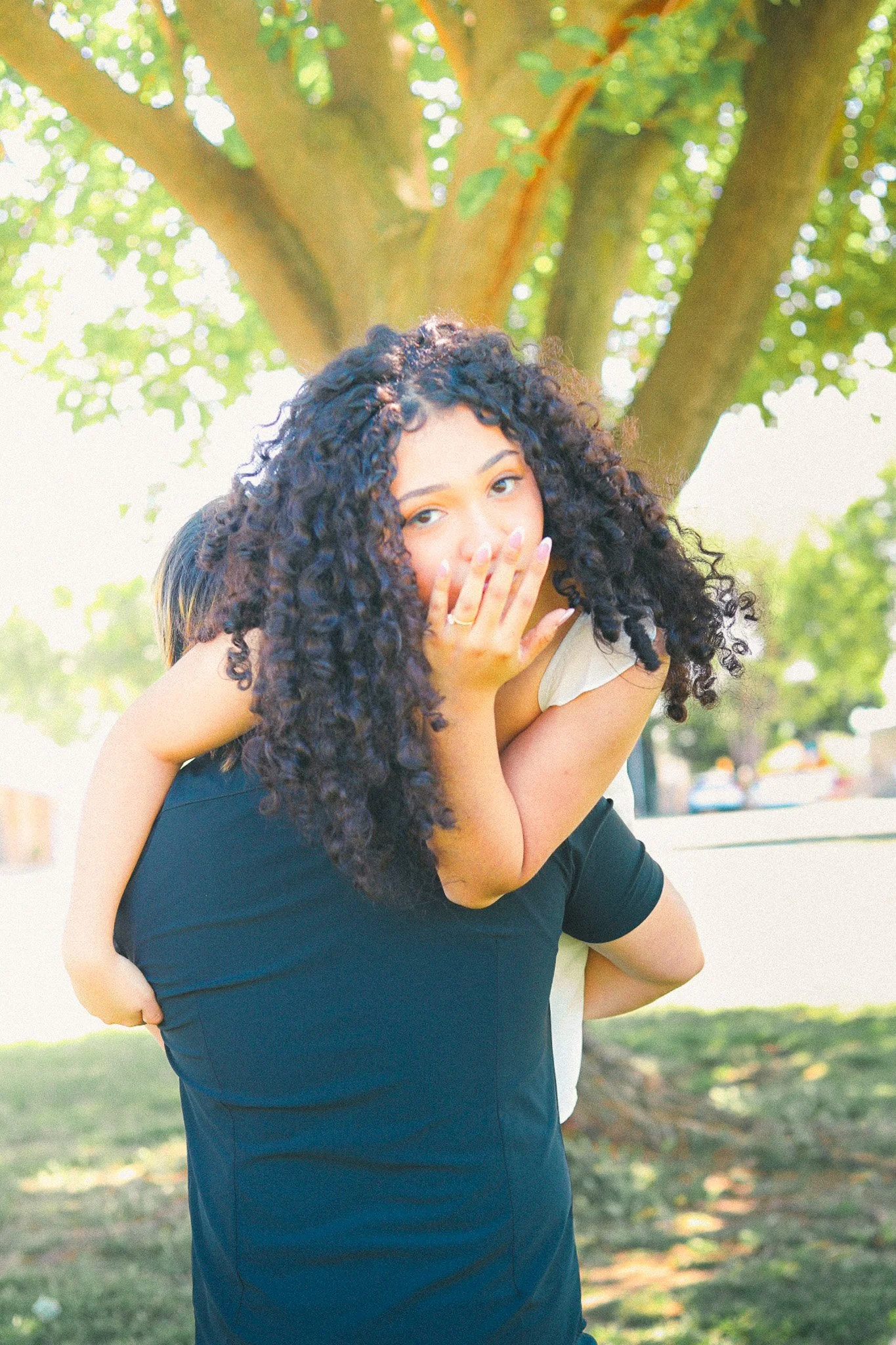 A young woman with curly hair being carried on a man's back outdoors under a large tree, covering her mouth with her hand and smiling.