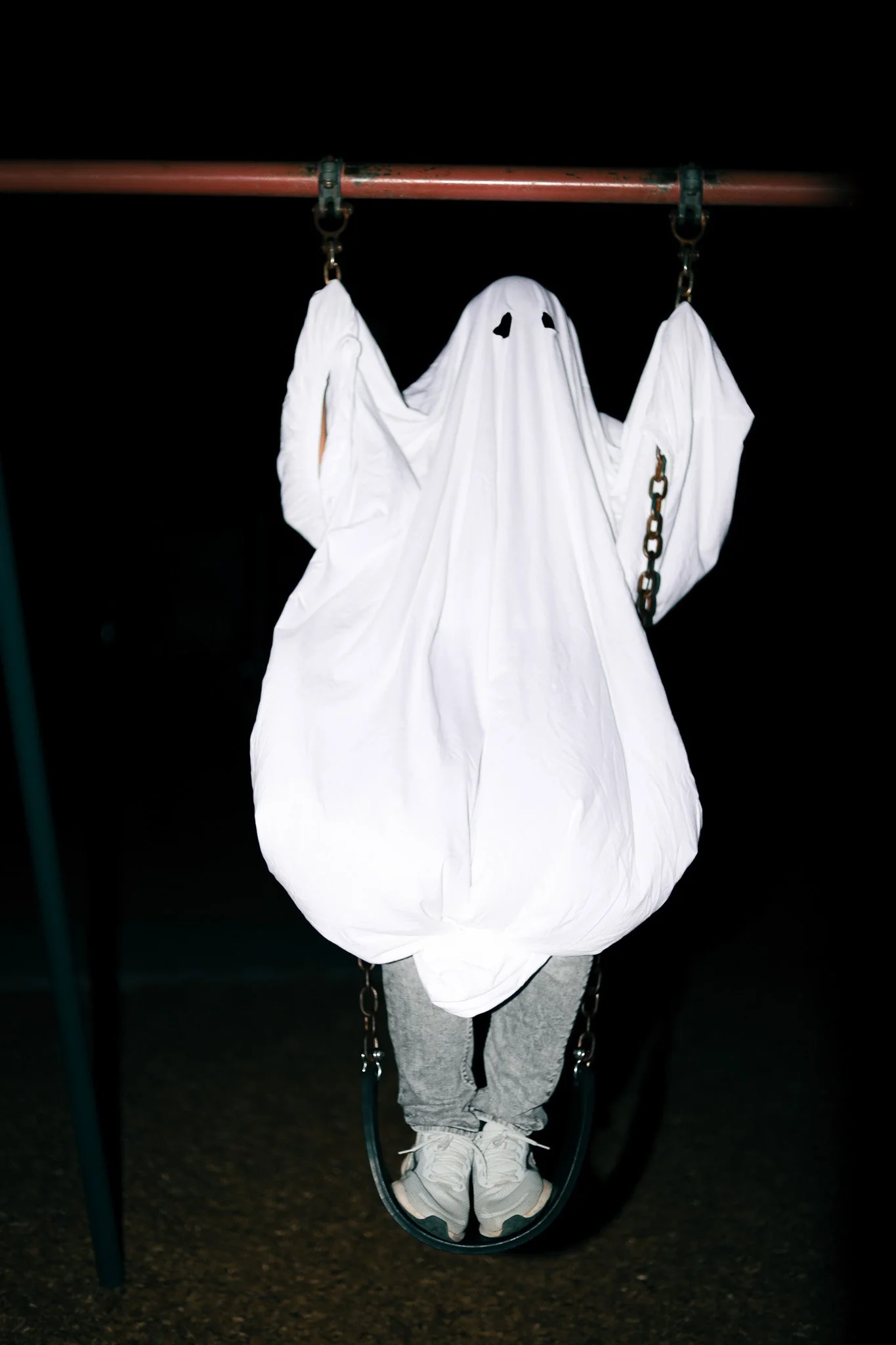 A swing set at night with a person dressed as a ghost wearing a white sheet with eye holes, hanging from the swing, with the person's legs and shoes visible beneath the sheet.