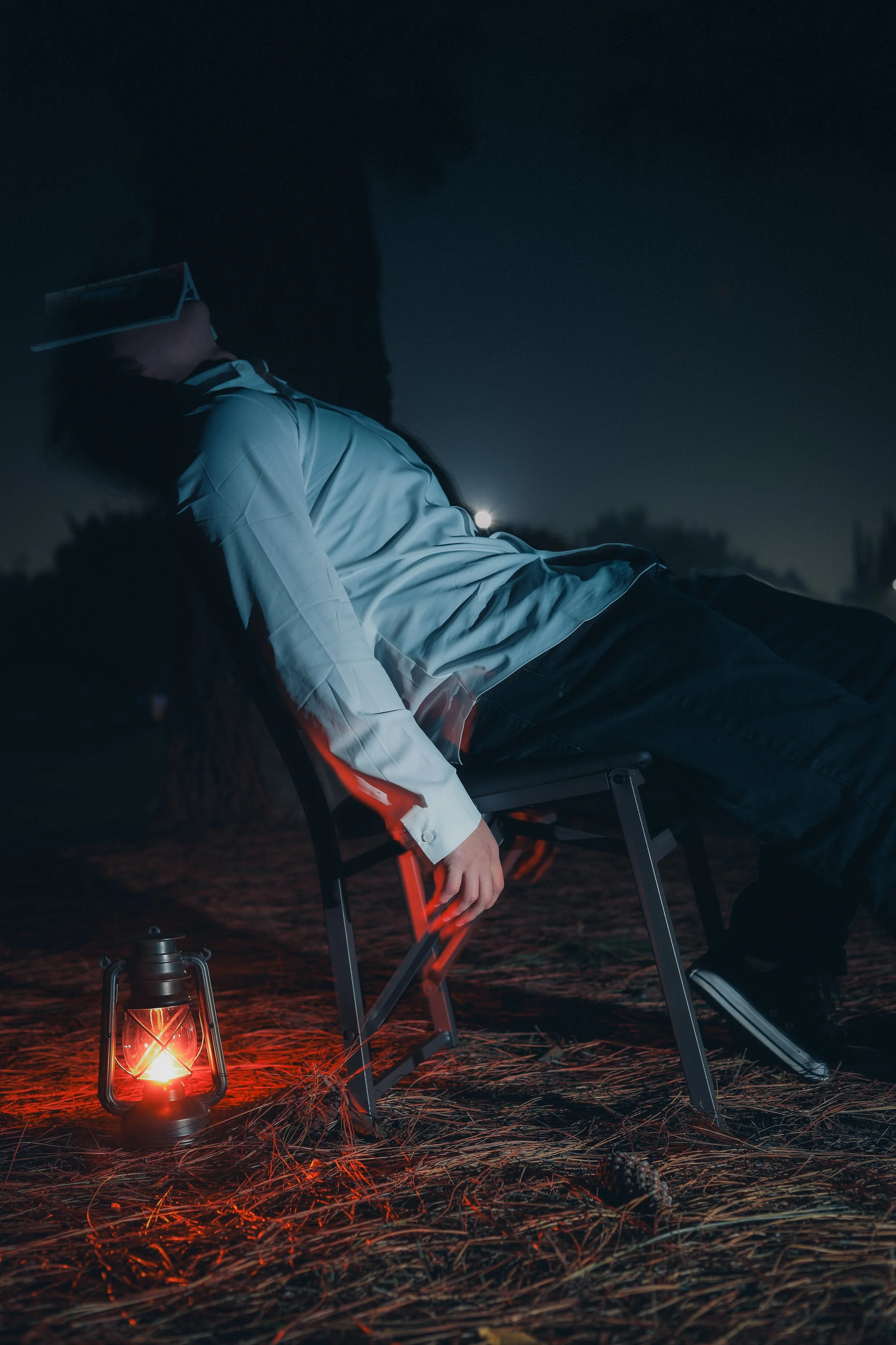 A person wearing glasses and a white shirt, reclining on a chair outdoors at night, with a lantern and pine cone on the ground and dark trees in the background.