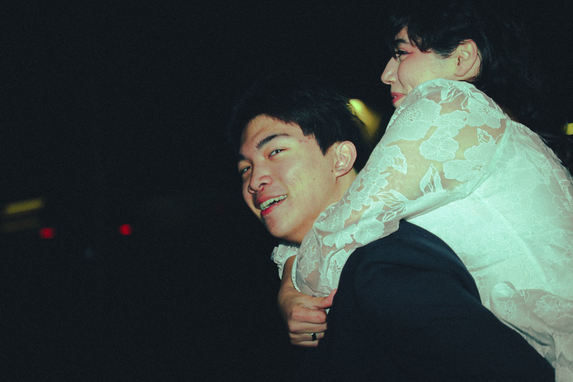 A young man giving a piggyback ride to a woman in a white floral lace top at a nightclub with dark background and red and yellow lights.