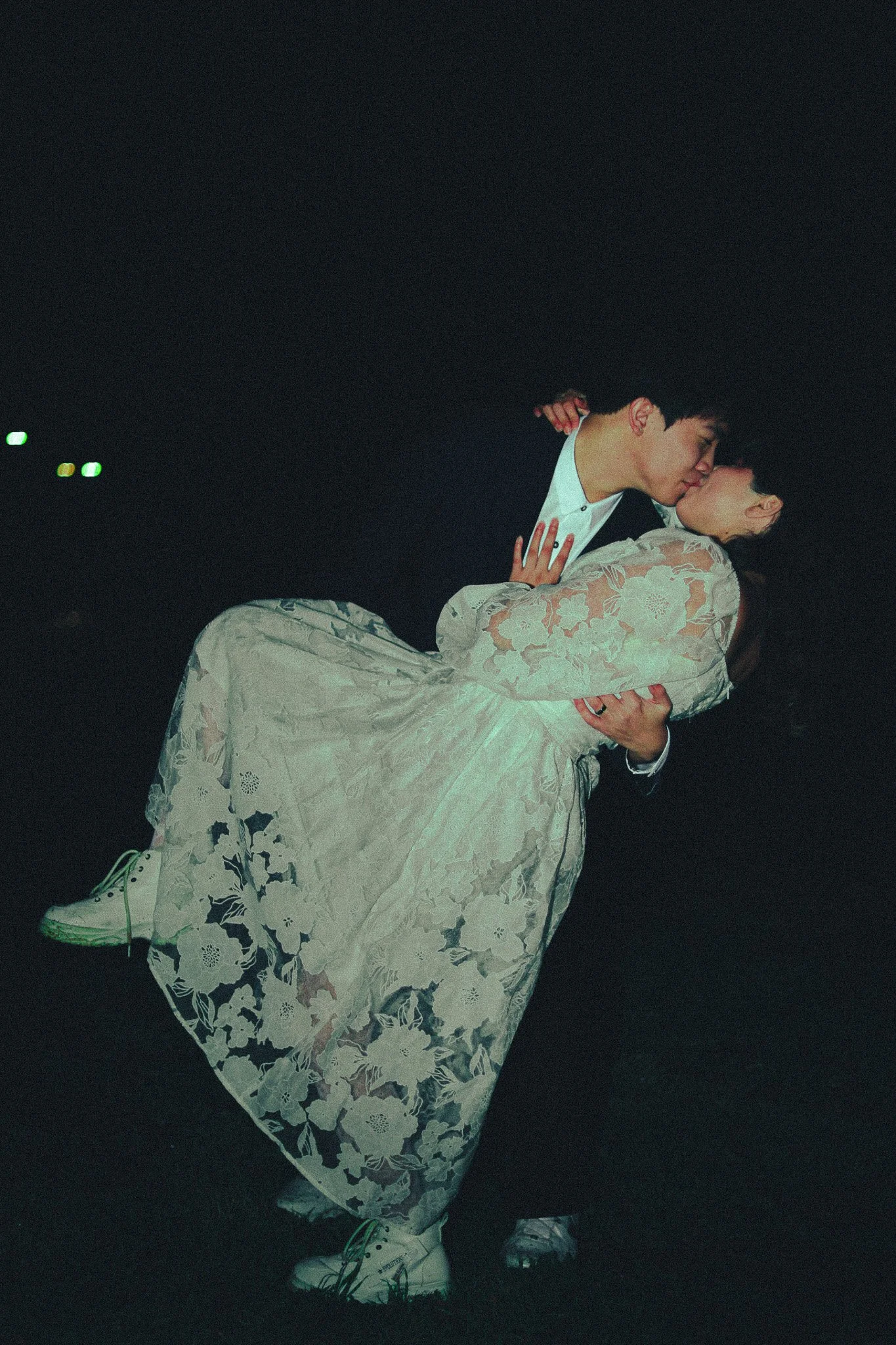 A couple sharing a kiss outdoors at night, with the man holding the woman in his arms. The woman is wearing a lace dress, and the man is dressed in a suit with white sneakers.