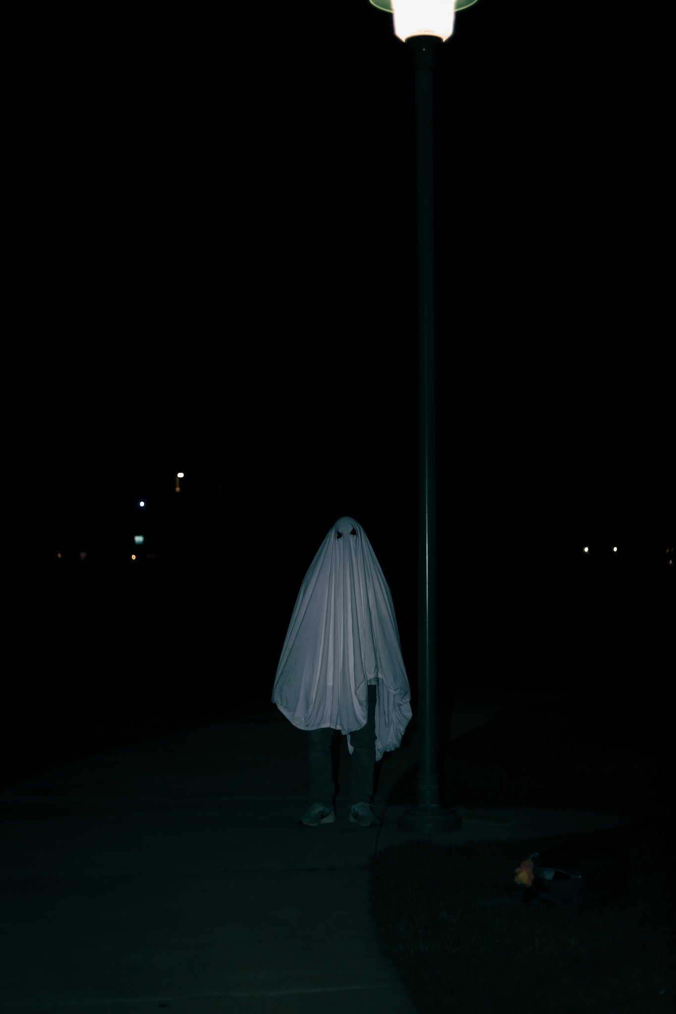 Person standing at night dressed as a ghost with a white sheet featuring eye holes, near a streetlamp on an empty street.