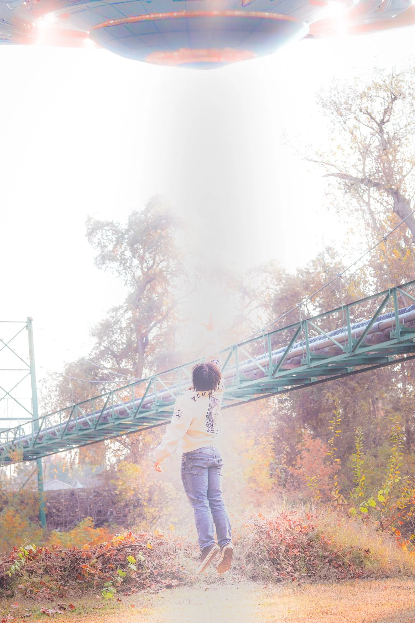 A person in a sweatshirt and jeans jumps on a grassy field during autumn. A large spaceship-like object is hovering above, and a metal bridge extends diagonally across the background. Trees with fall foliage surround the scene.