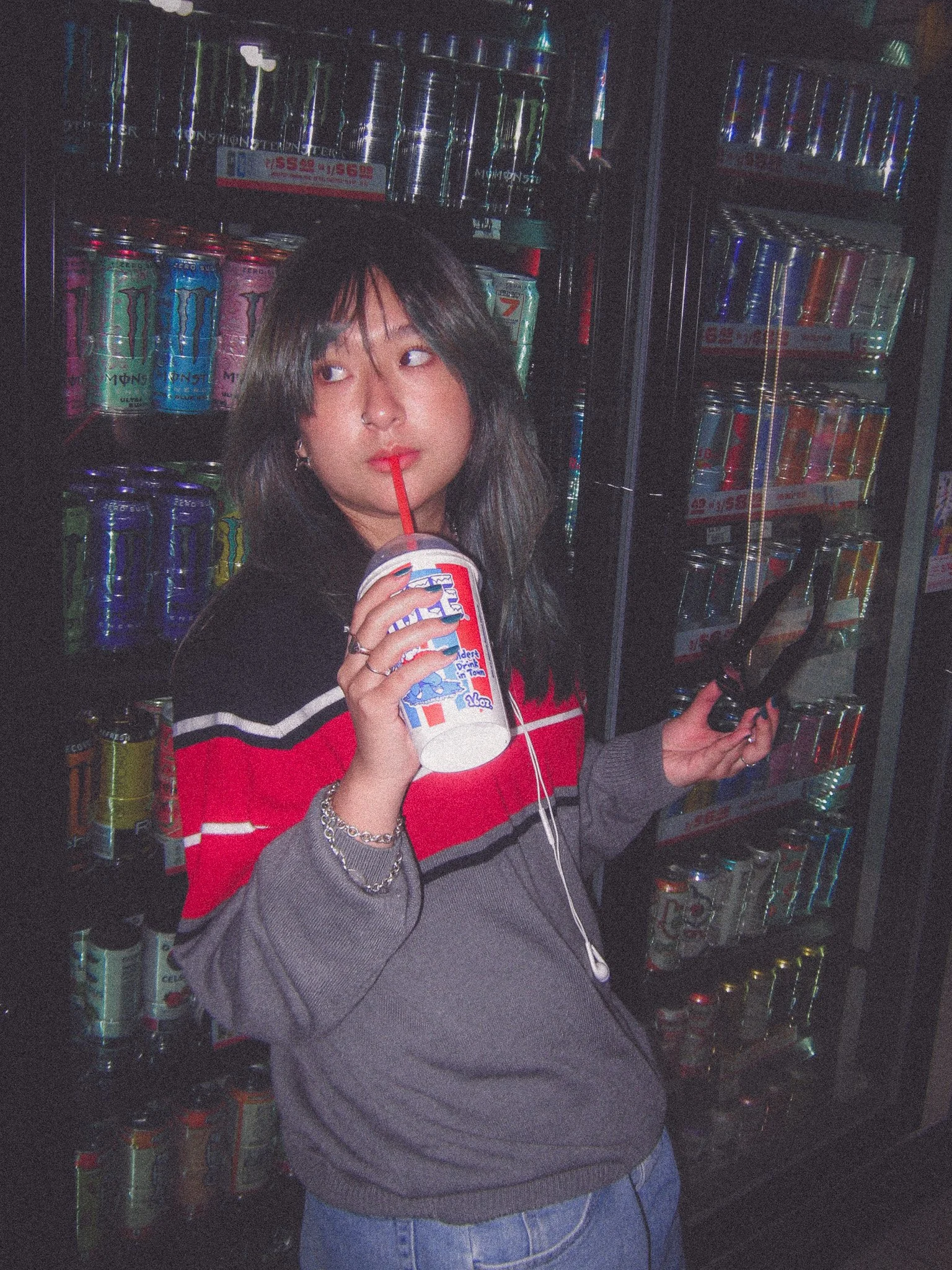 A young woman with dark hair and light skin sipping a drink through a red straw from a fast-food cup in front of a vending machine filled with canned beverages at night.