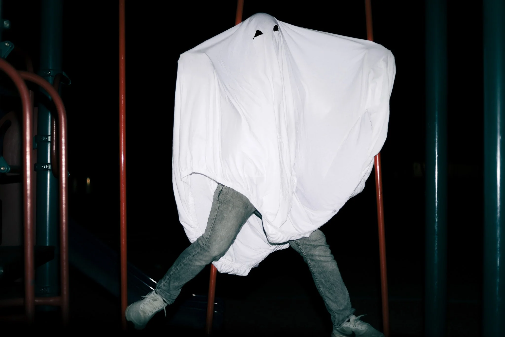 A person with jeans and sneakers, partially covered by a white sheet, walking on playground equipment at night.