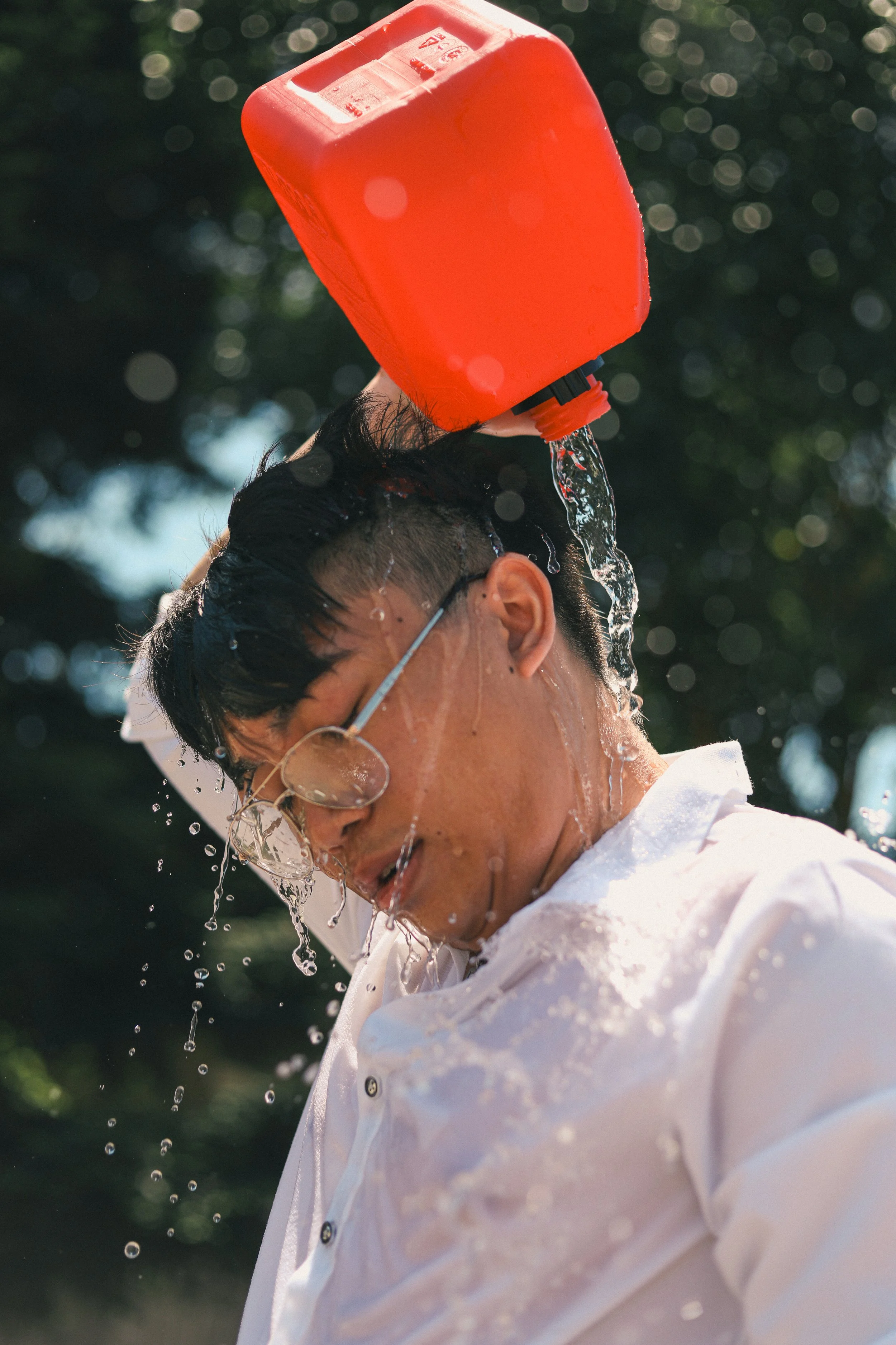 Man with glasses pouring water over his head from a red container.