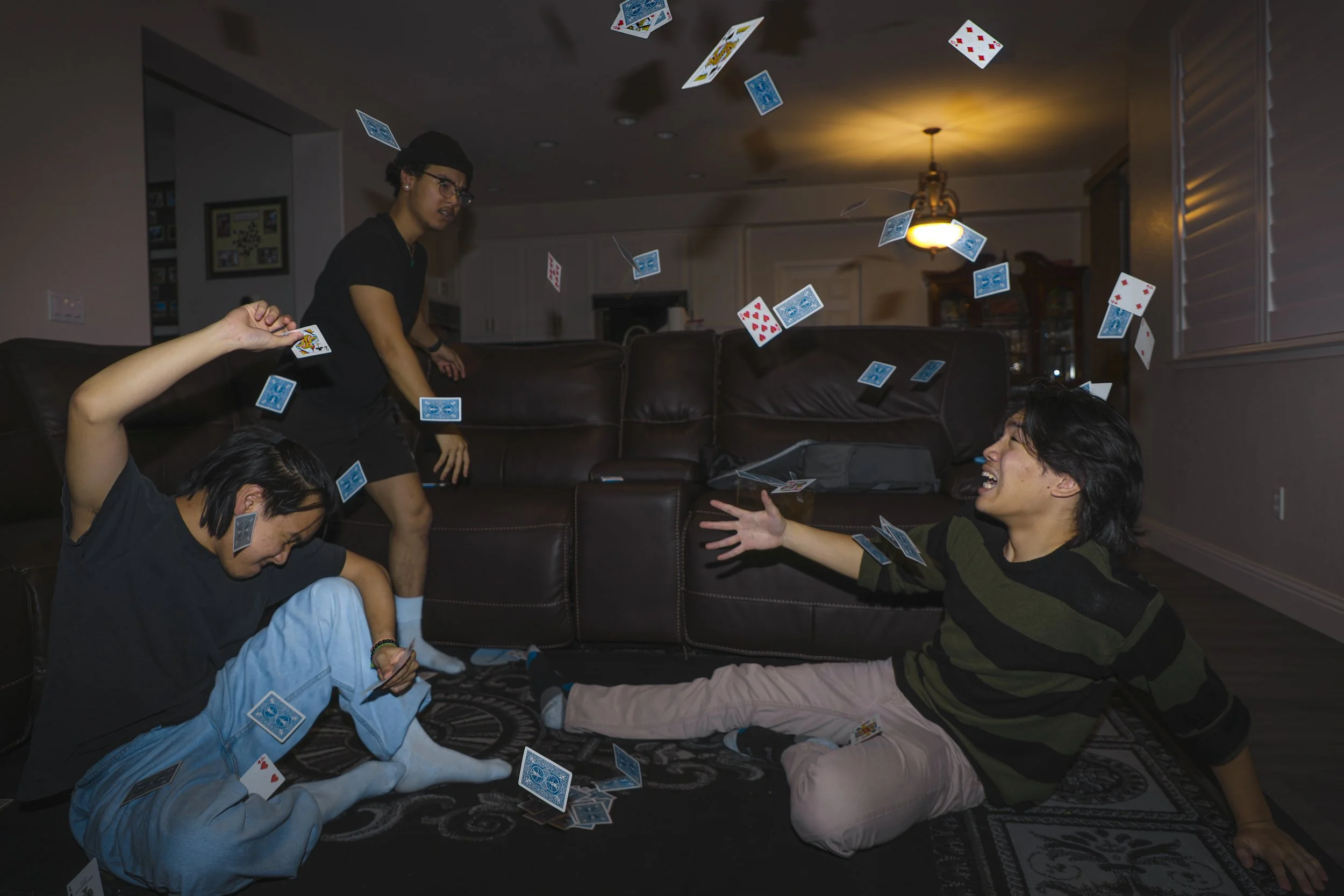 Four young people playing cards indoors, with cards flying around in the air. Two women are seated on the floor, one holding cards and the other laughing with arms outstretched, while two men are standing or sitting nearby.