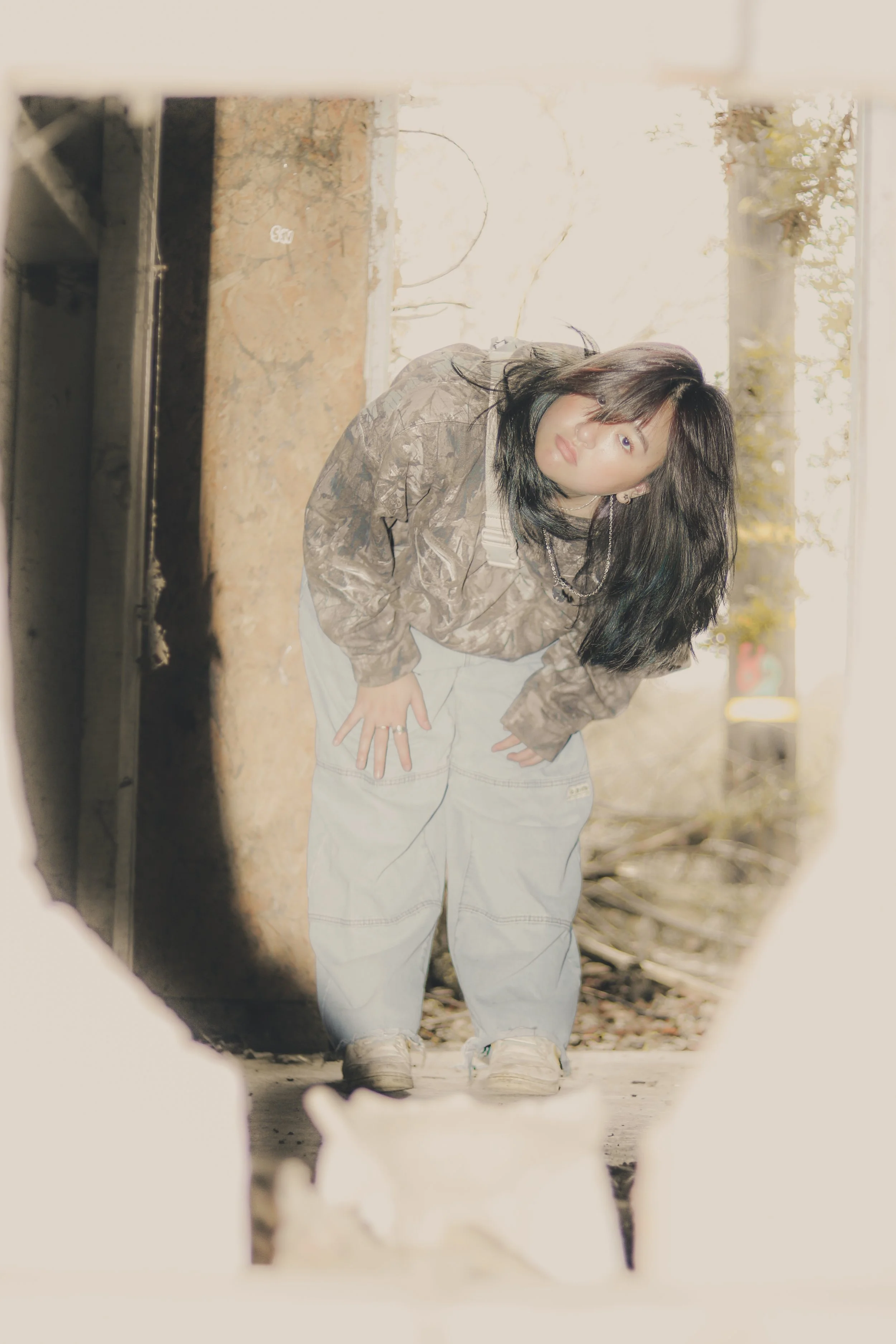 Woman with black hair in a camouflaged jacket and light-colored jeans leaning forward, viewed through a hole or opening in a surface, with sunlight and trees in the background.