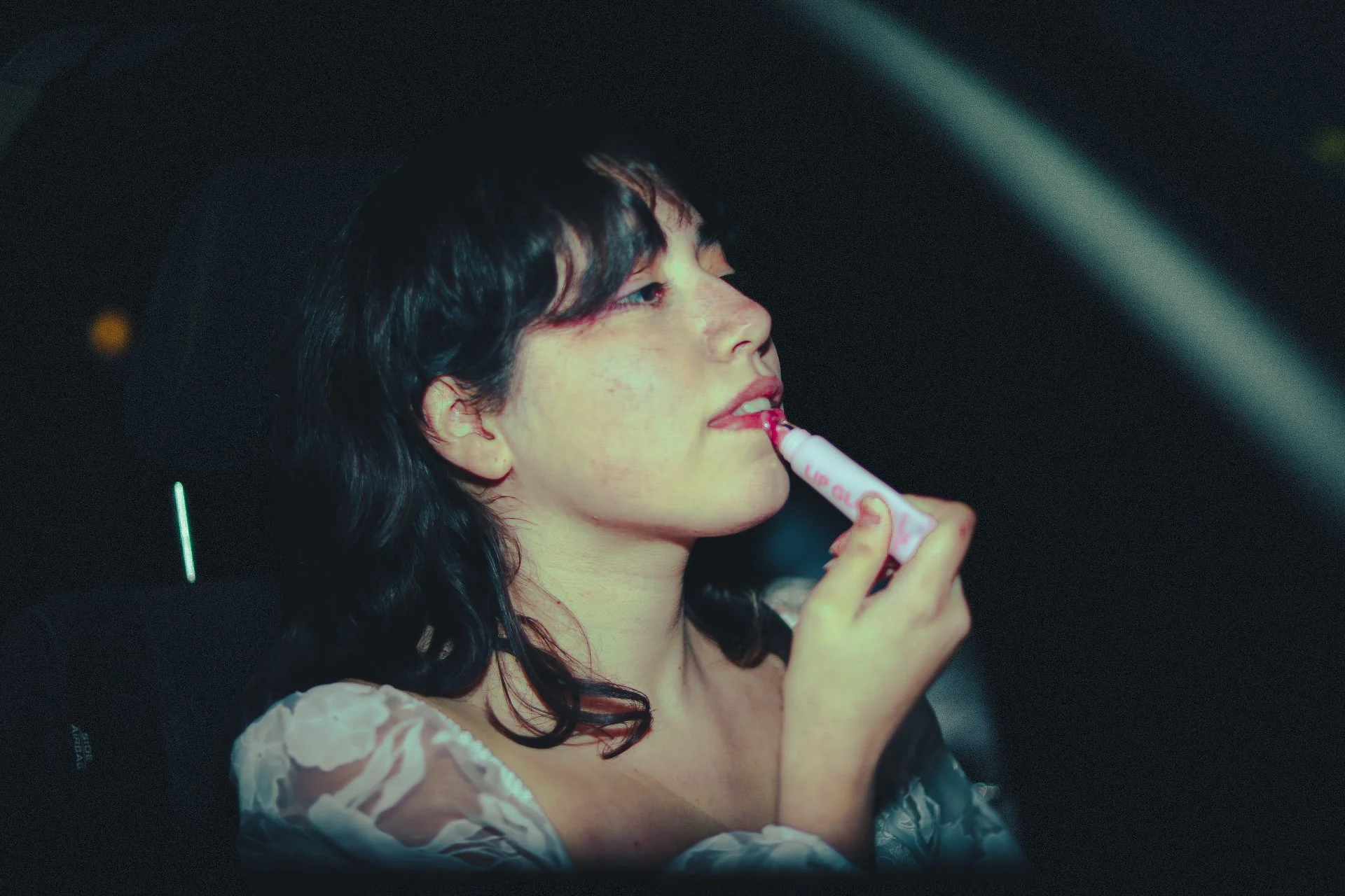 A young woman with black, wavy hair applying red lipstick in a dark environment.