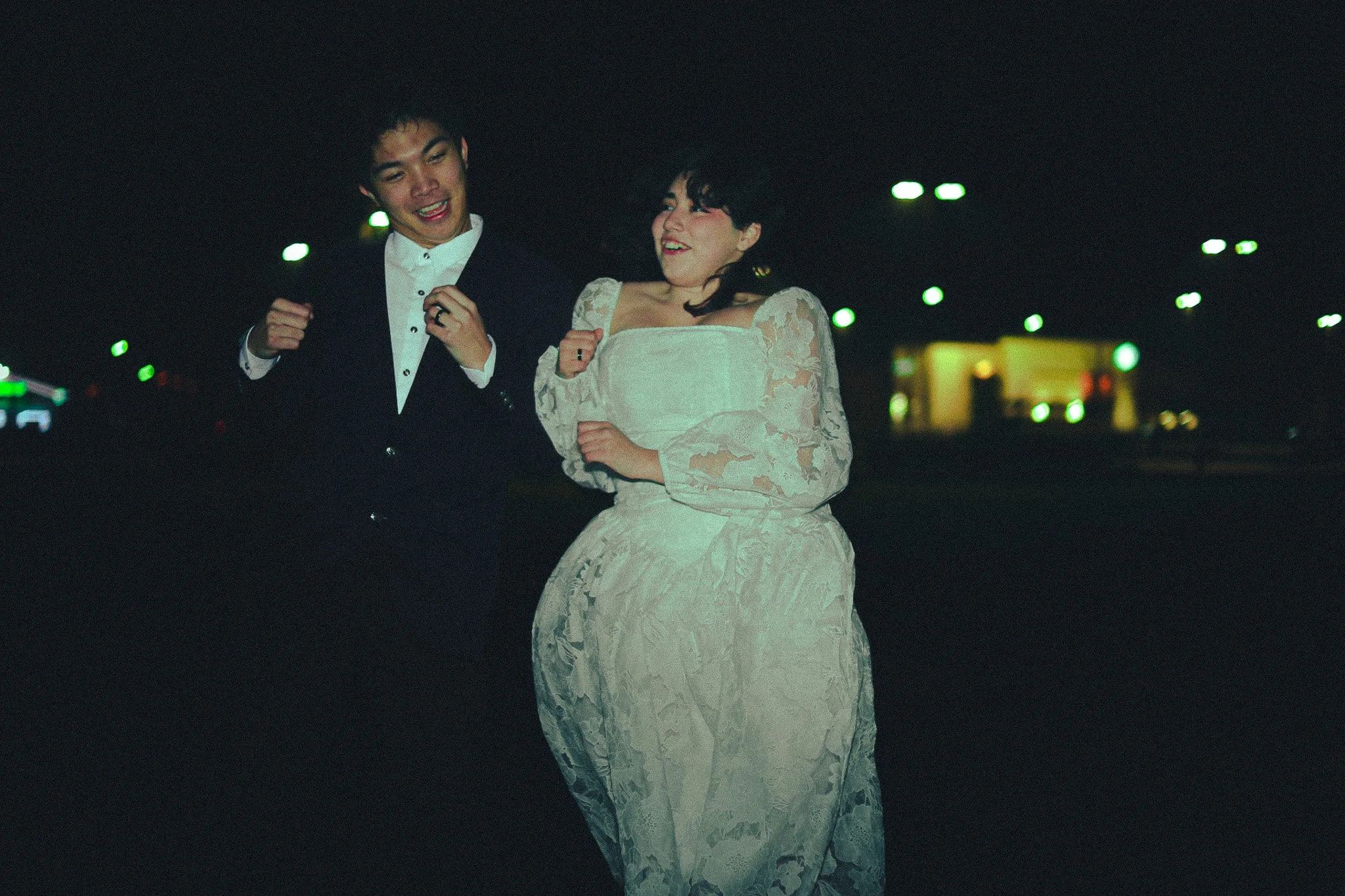 A man and woman dressed in formal attire, walking together at night, with the woman wearing a white lace dress and the man in a black tuxedo.