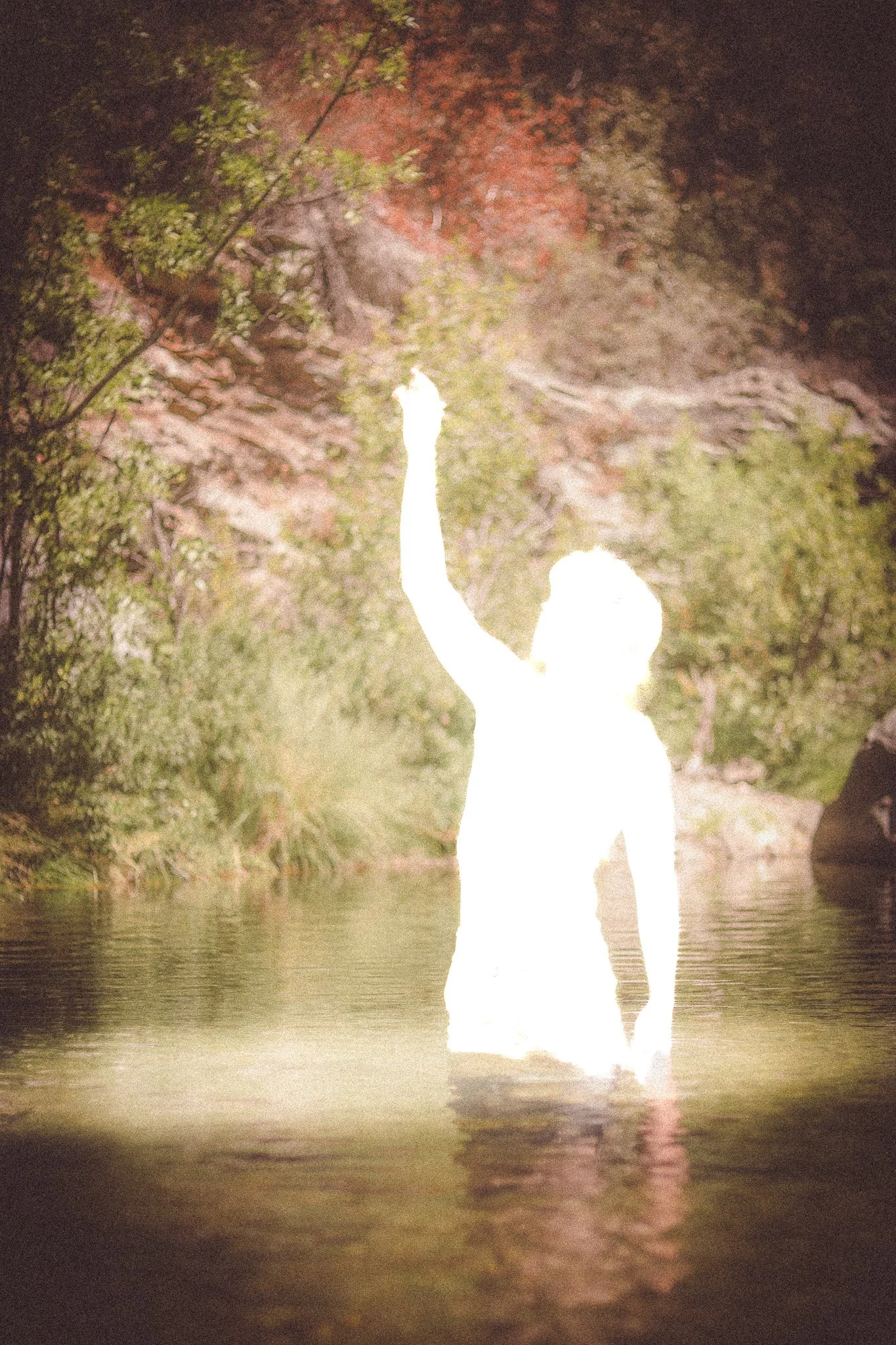 A person standing waist-deep in water, raising one arm, surrounded by trees and mountains.