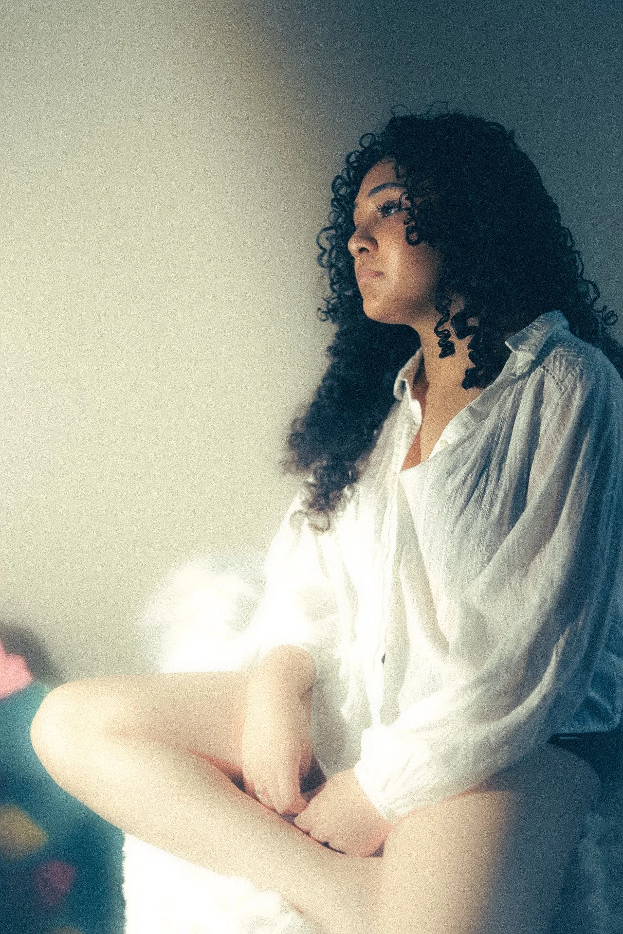 A woman with long curly black hair, wearing a white shirt, sitting with her legs crossed in a room with plain walls, illuminated by soft natural light.