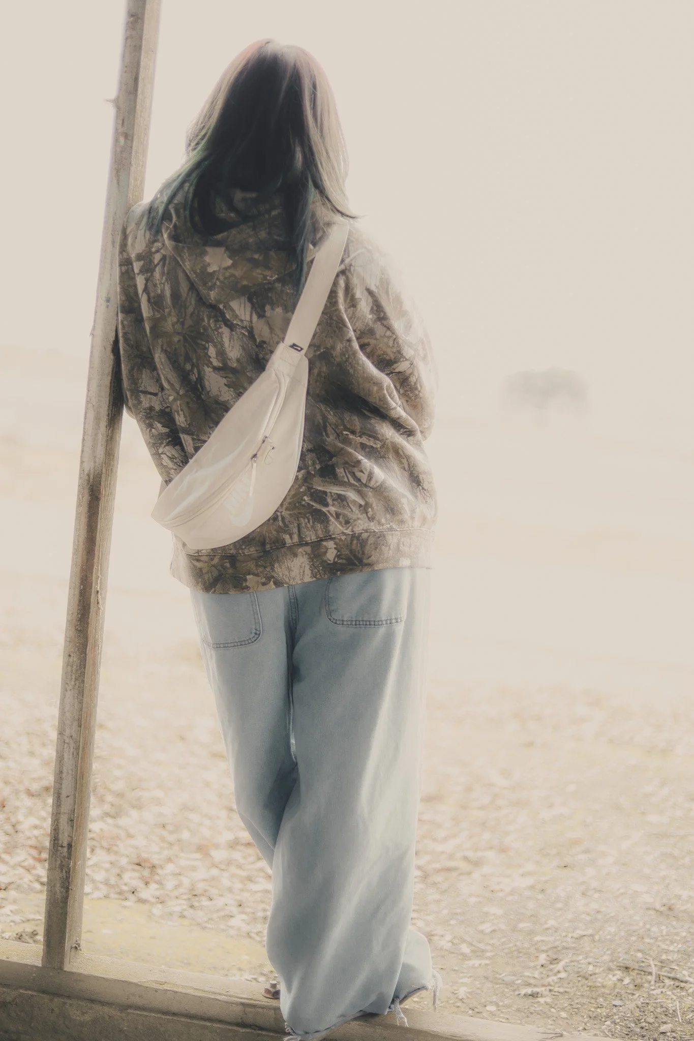 Person standing outdoors near a wooden post, facing away, wearing a camouflage jacket, light blue jeans, and carrying a light-colored shoulder bag.