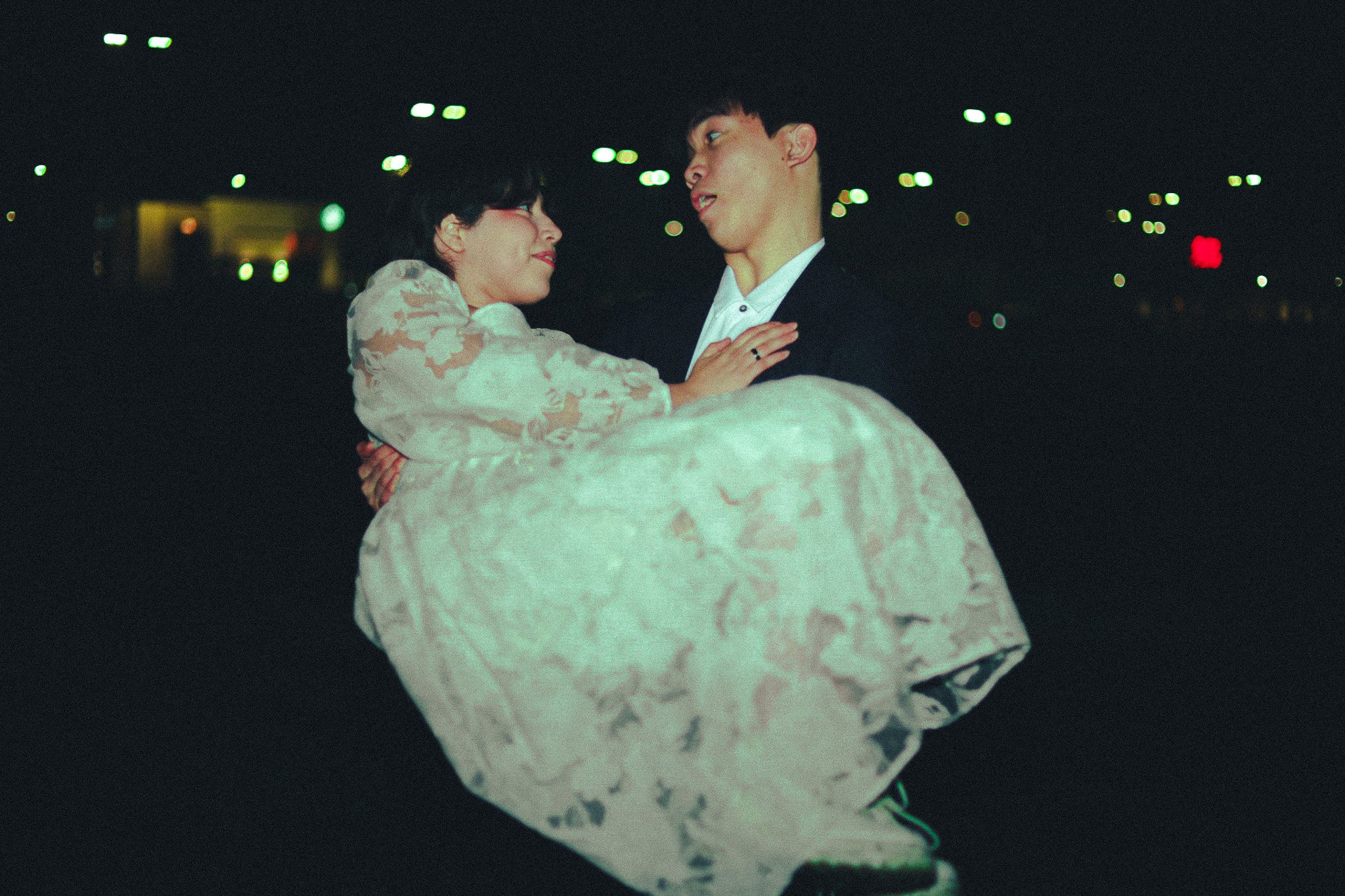 A young man in a dark suit is holding a woman in a light dress in his arms in a dark, urban setting at night, with city lights in the background.