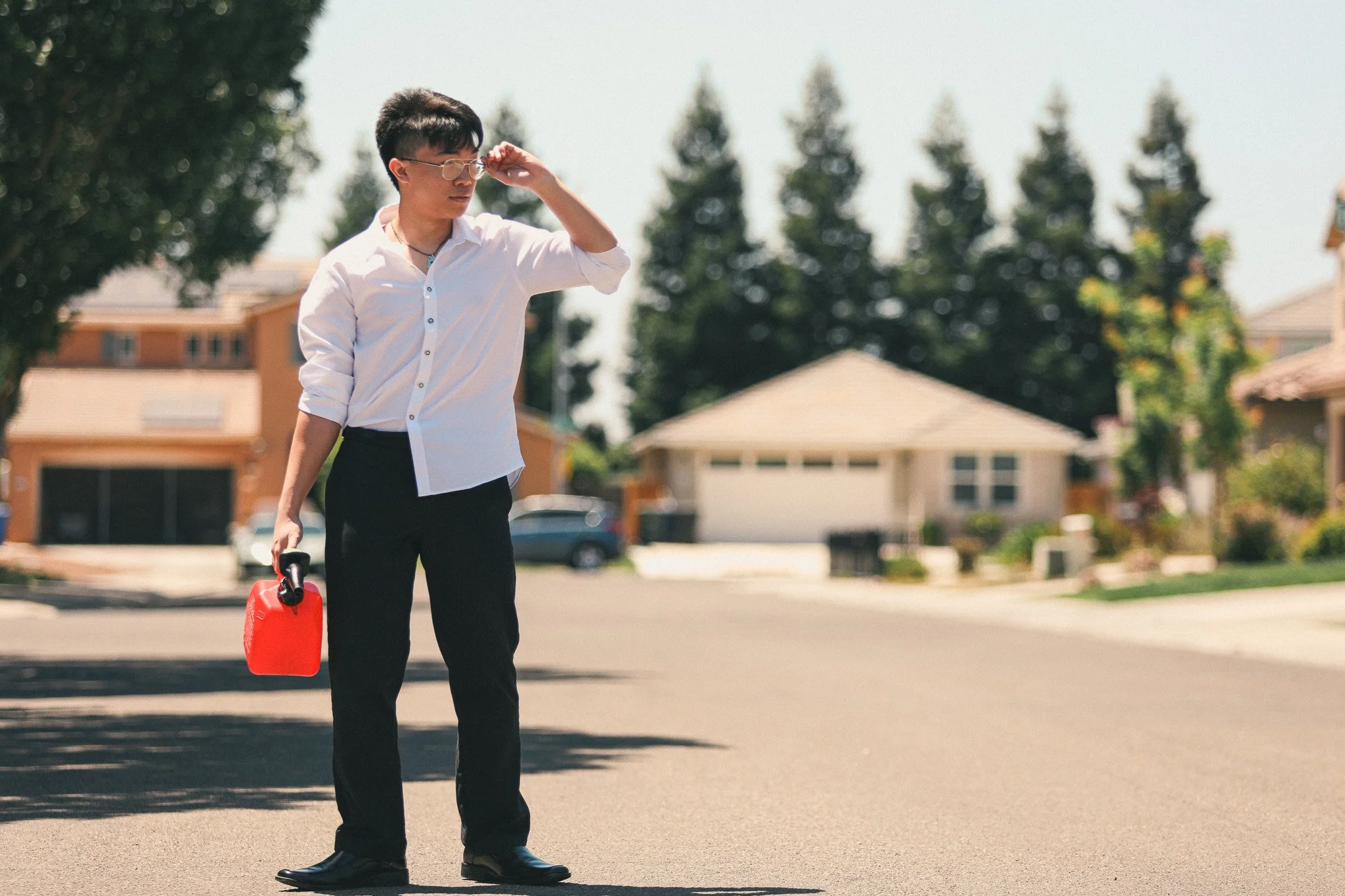 A young man in business attire walking on a suburban street holding a red gasoline can, adjusting his glasses with a cityscape in the background.