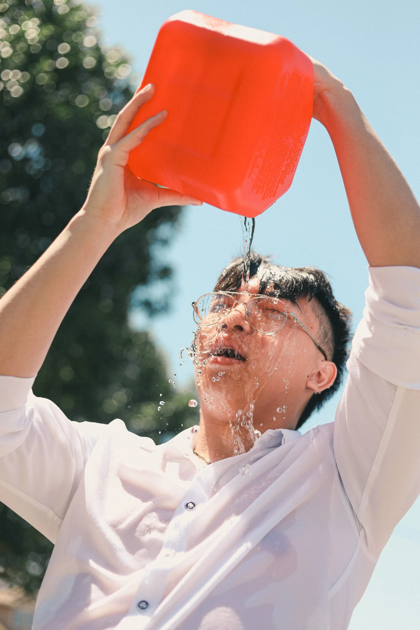 Person with short hair and glasses pouring water from a large red container outdoors on a sunny day.