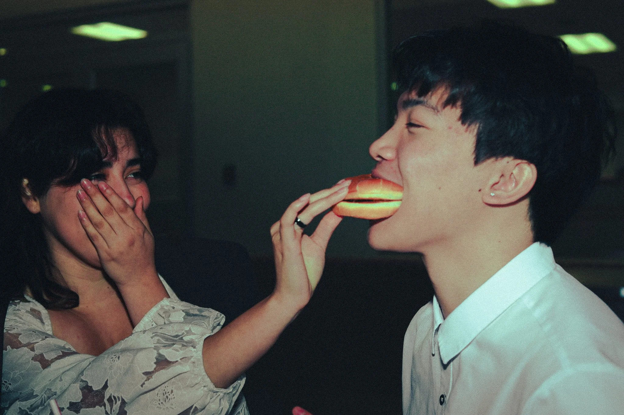 A woman covering her mouth in surprise while a young man with tattoos and earrings is about to take a bite of a burger