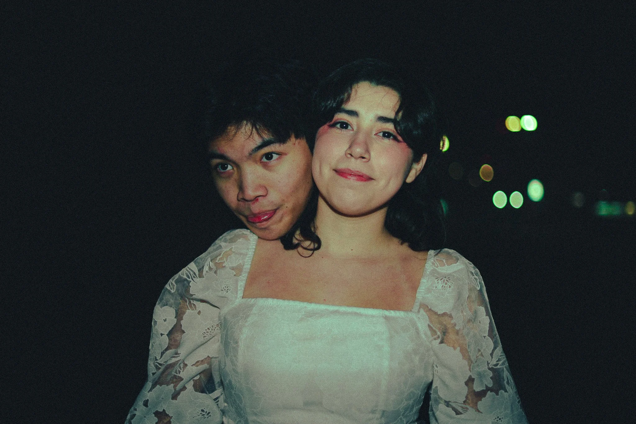 A young man and woman posing together at night, with the man playfully sticking his tongue out behind the woman's shoulder. The woman is smiling softly and wearing a white lace dress.