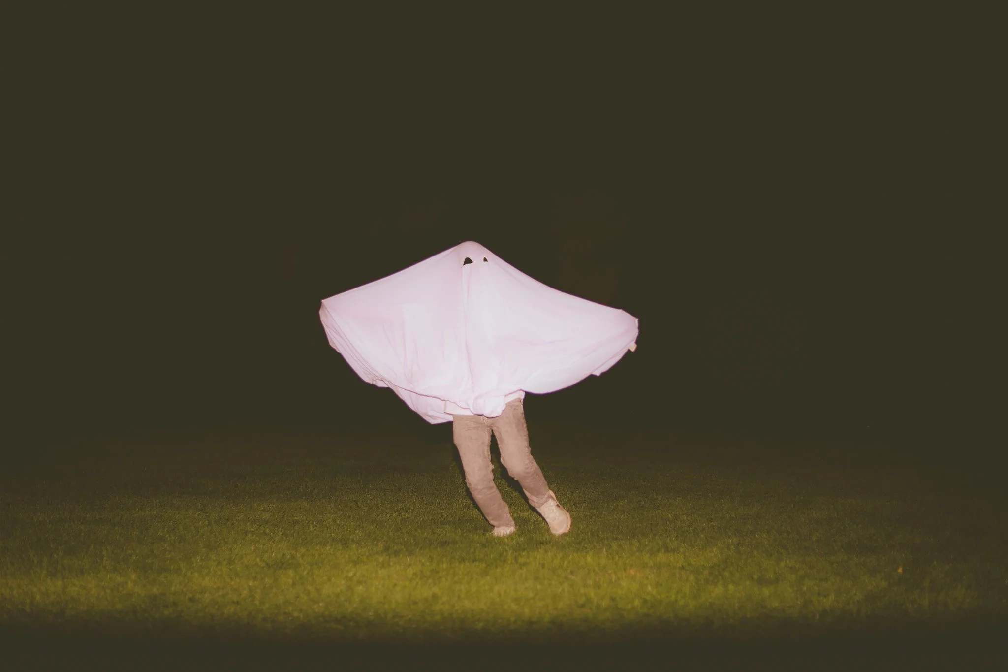 Person outdoors at night wearing a cape that resembles a ghost or a sheet, with a pink glow, and standing on grass