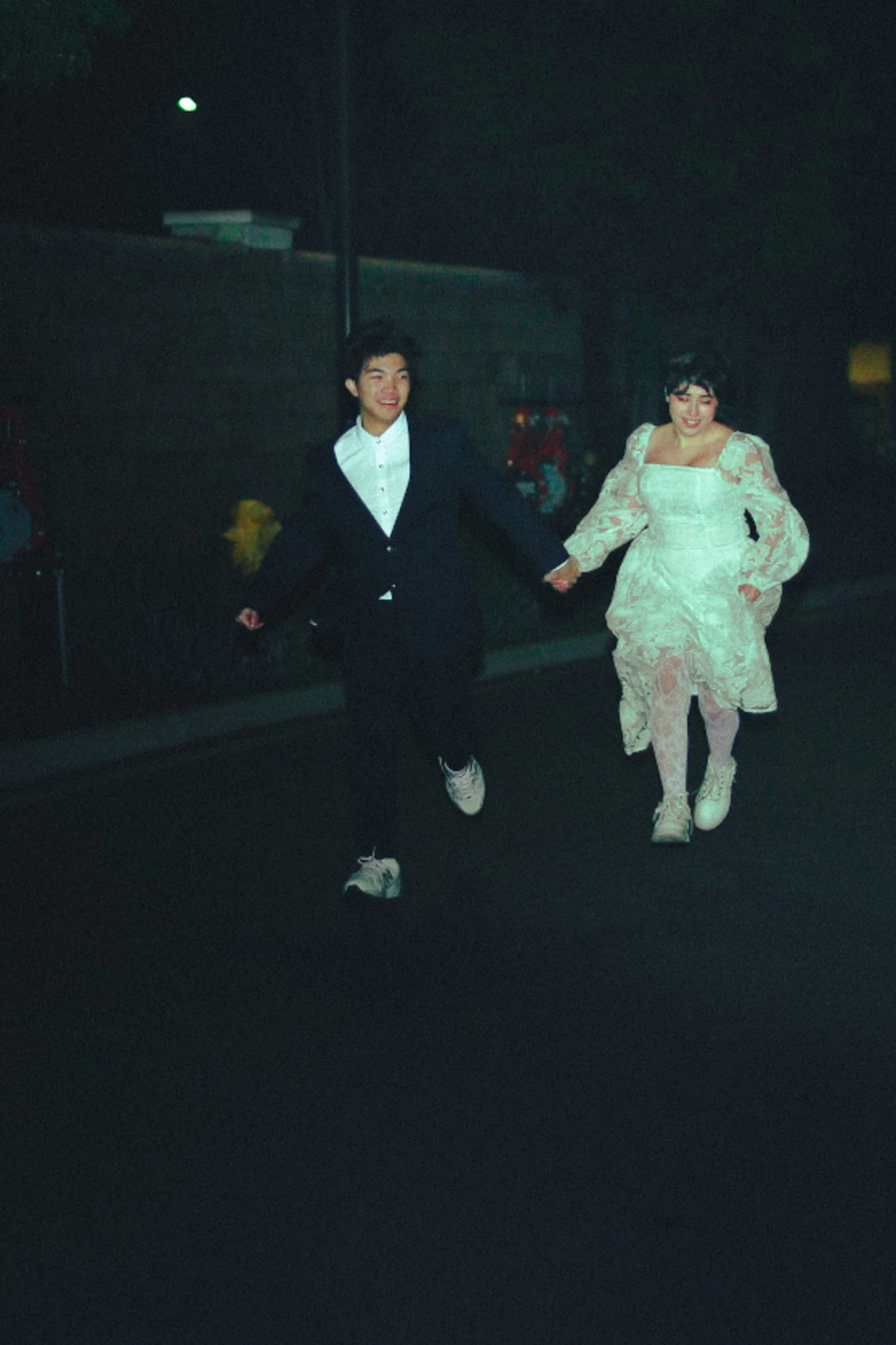 Two people holding hands and running outdoors at night, dressed in formal attire, smiling.