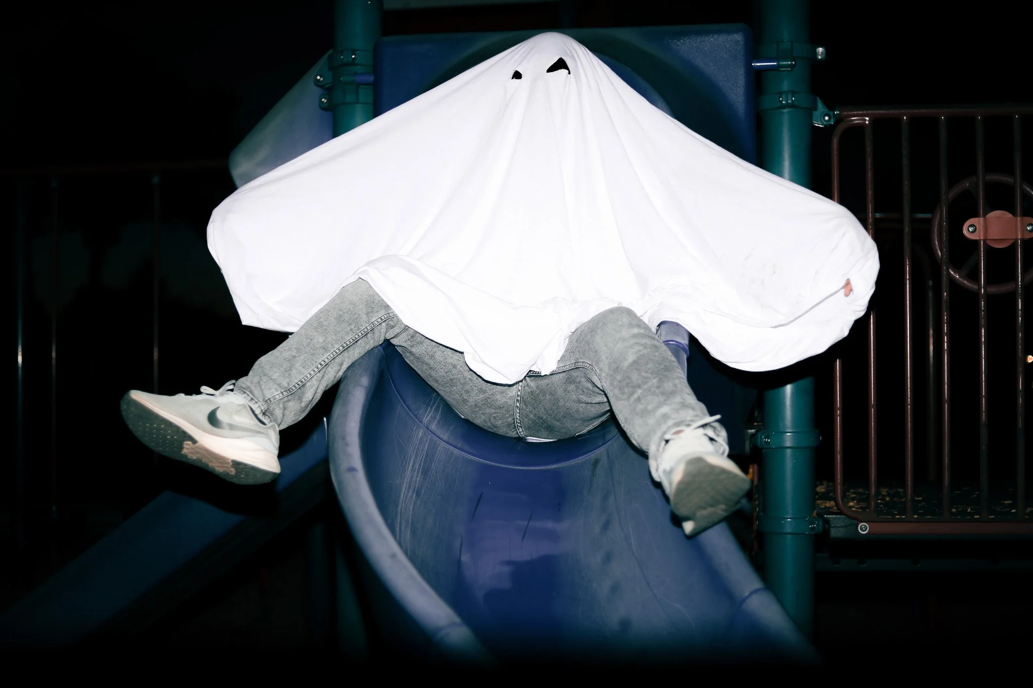 Child wearing a white sheet as a ghost costume, sitting at the top of a playground slide at night.
